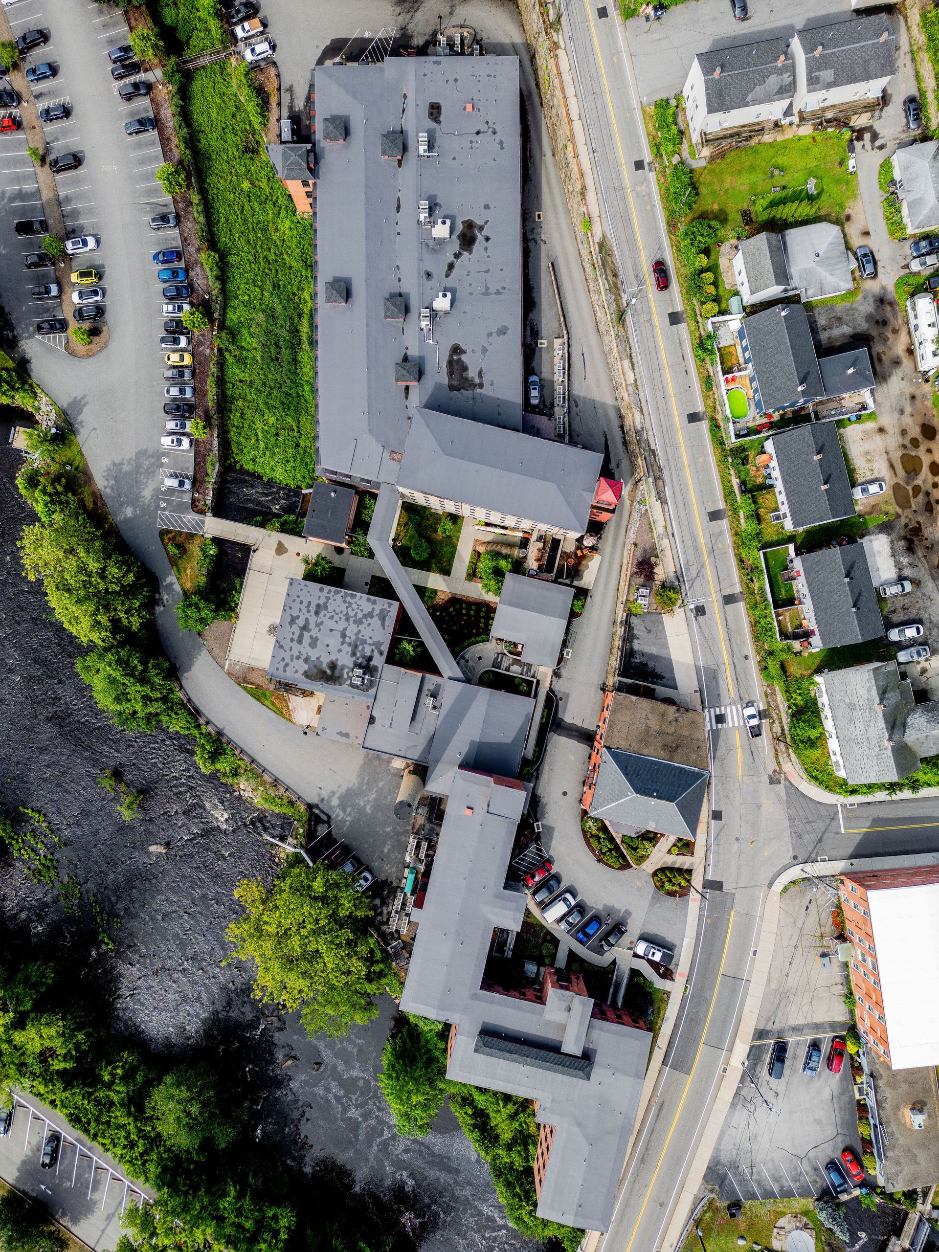 Aerial view of a building complex beside a road and parking lot, surrounded by trees and nearby houses.