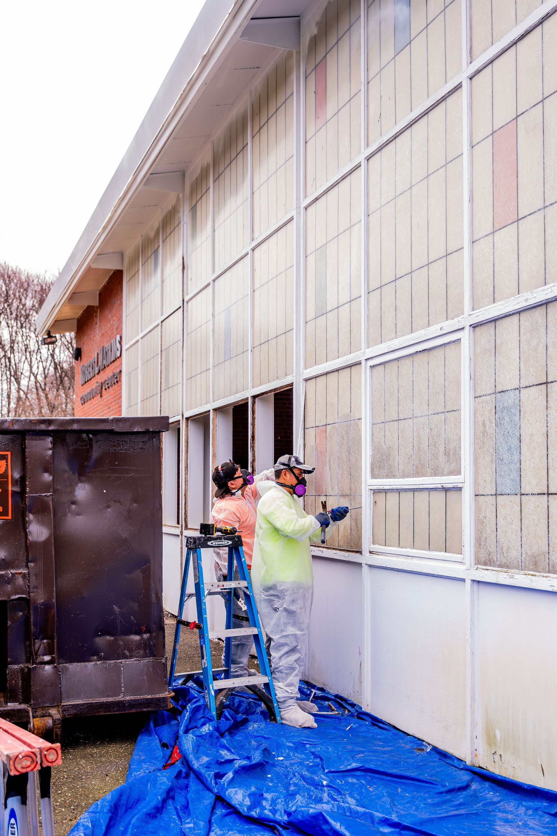 Two workers painting an exterior wall on blue tarp, one on a ladder beside a black dumpster.