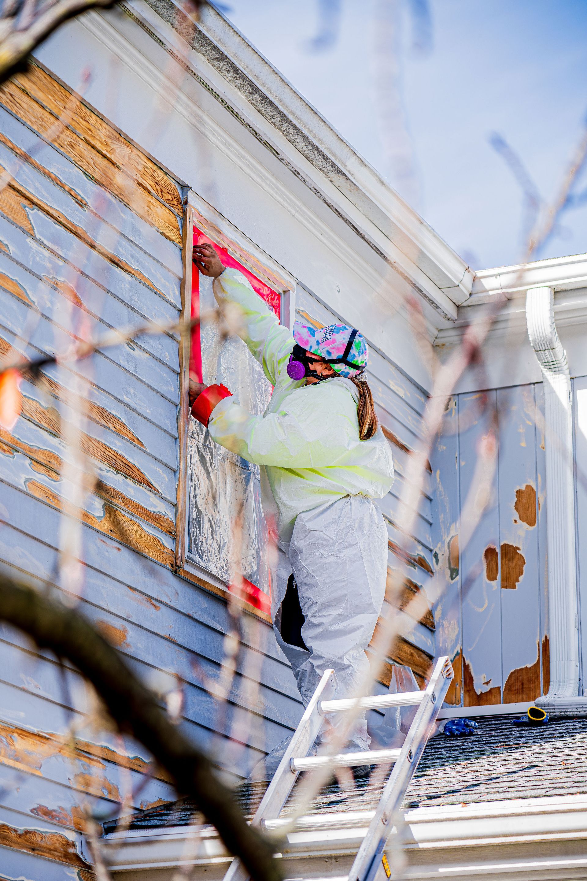Worker painting a house exterior on a ladder, using a roller on light siding with red trim.