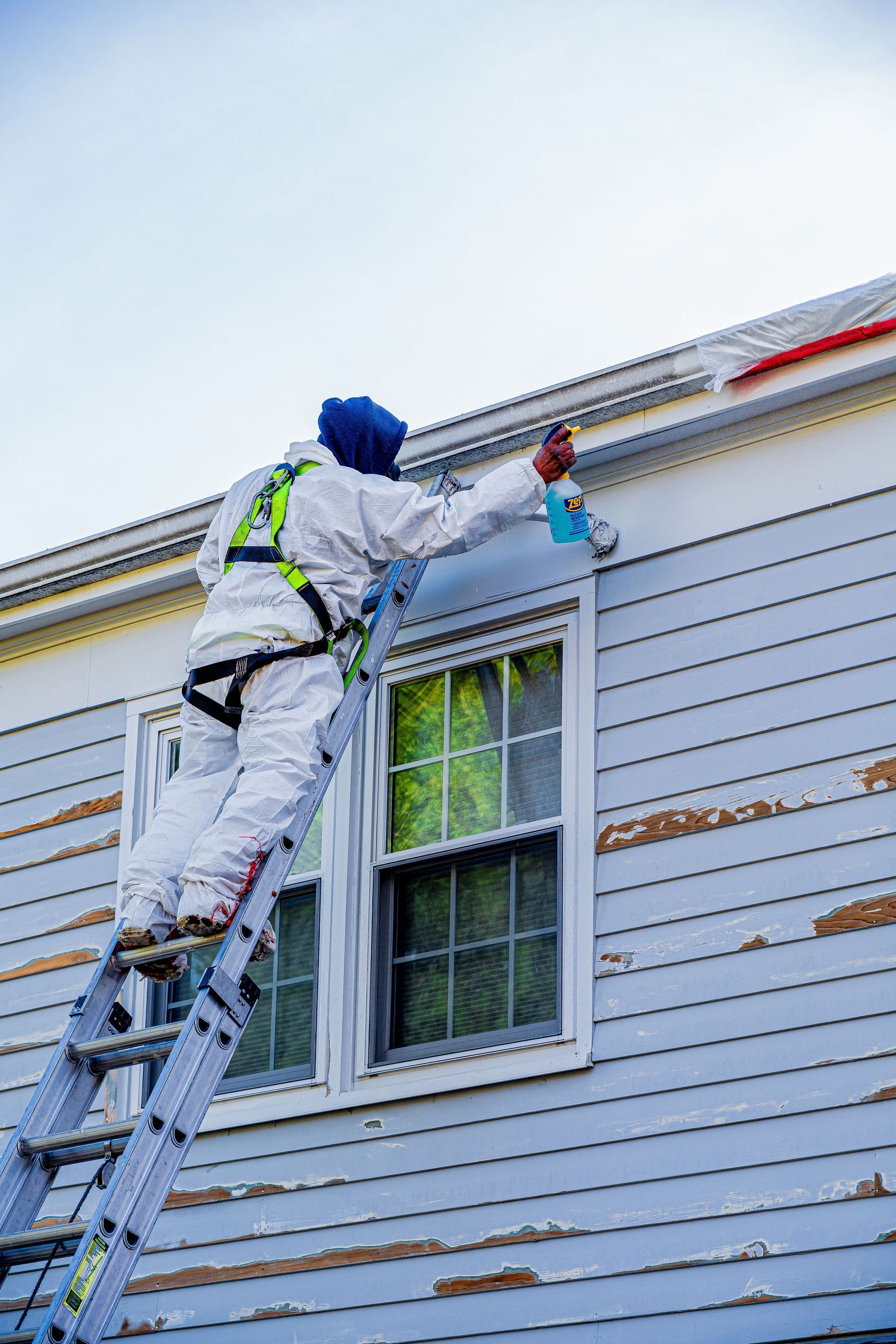Worker on a ladder painting a white house exterior near the roofline.