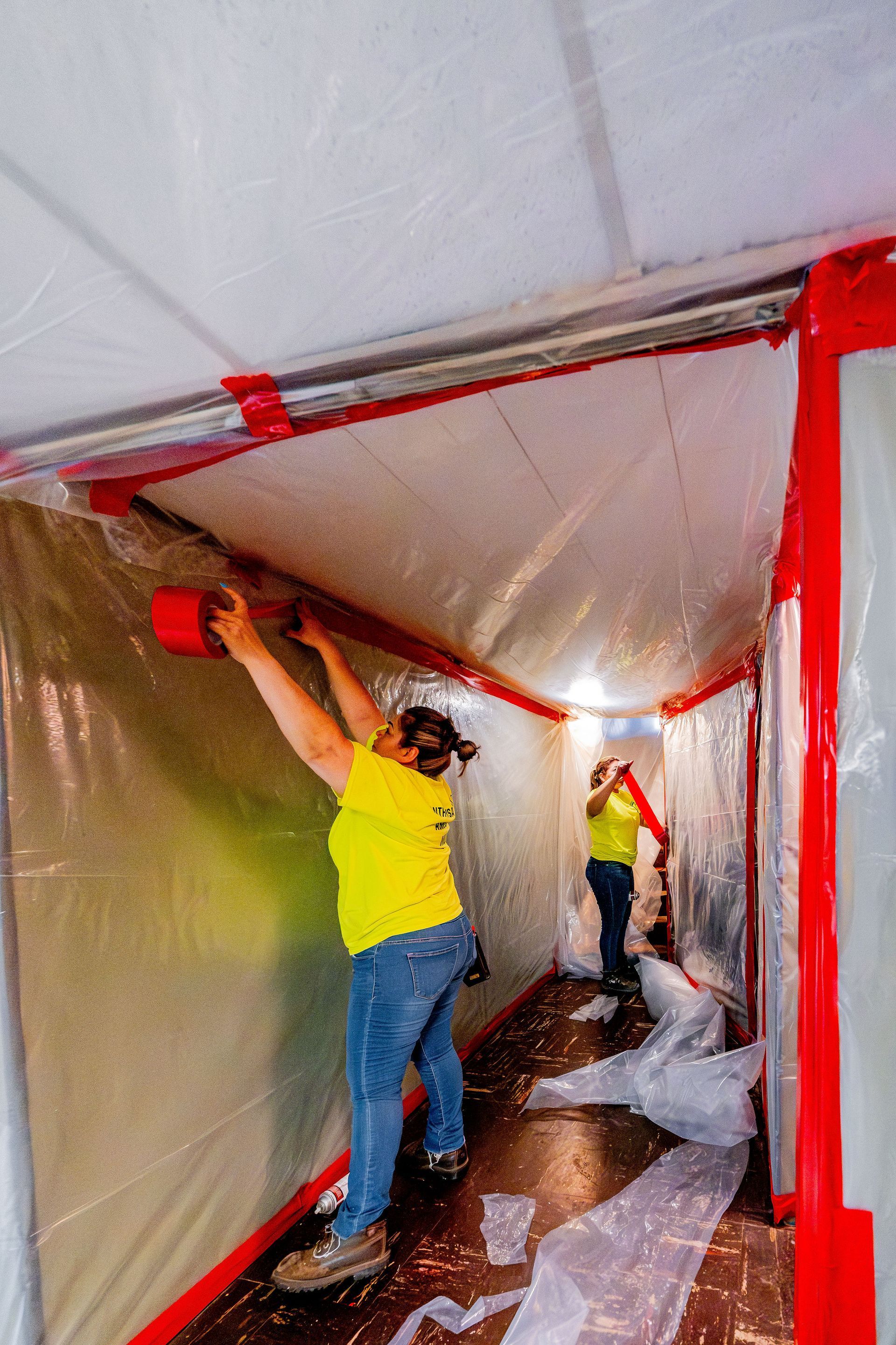 Two workers in yellow vests tape plastic sheeting inside a narrow hallway during renovation.