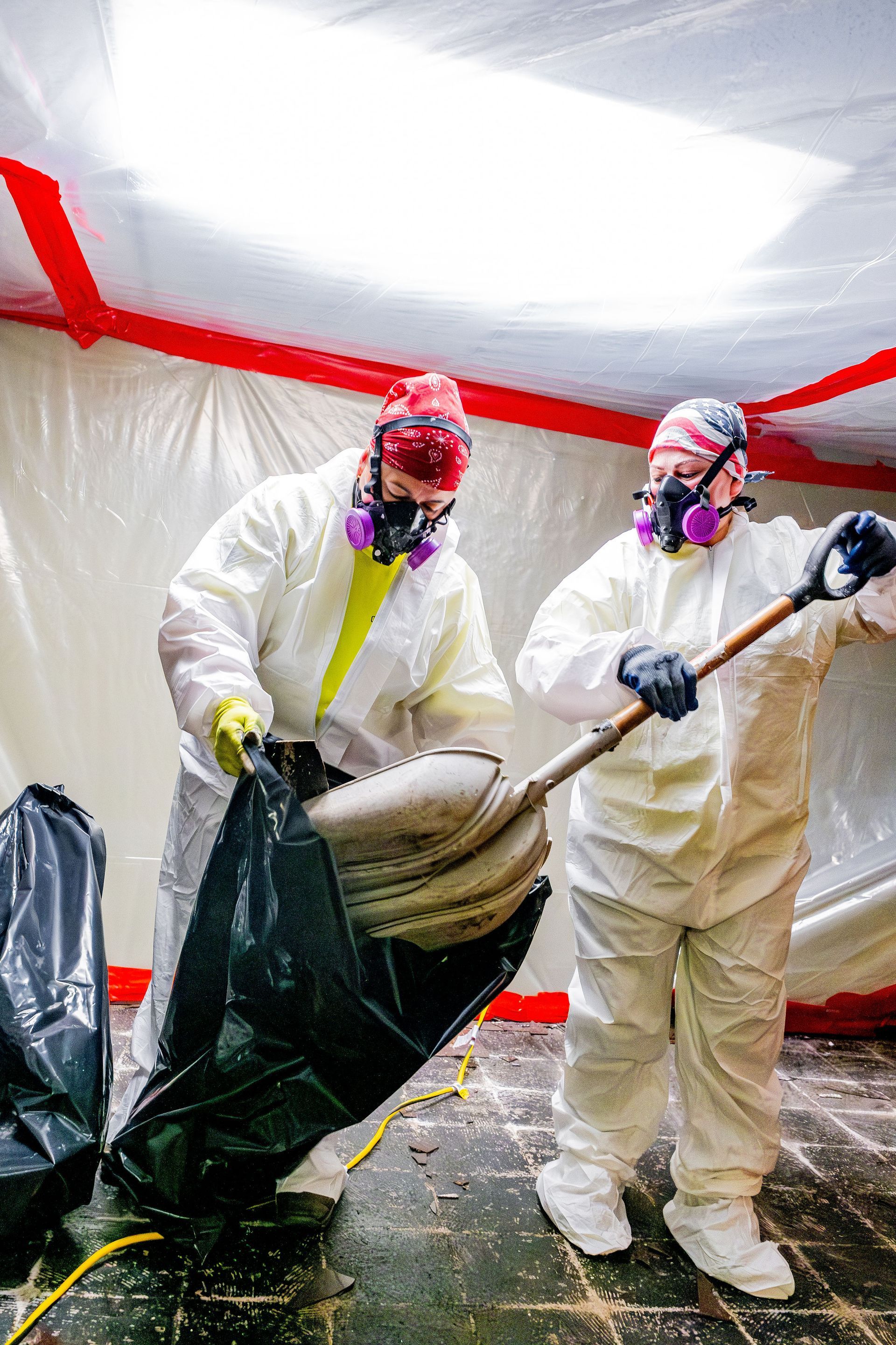 Two people in hazmat suits handling a black bag inside a plastic-lined contamination tent.