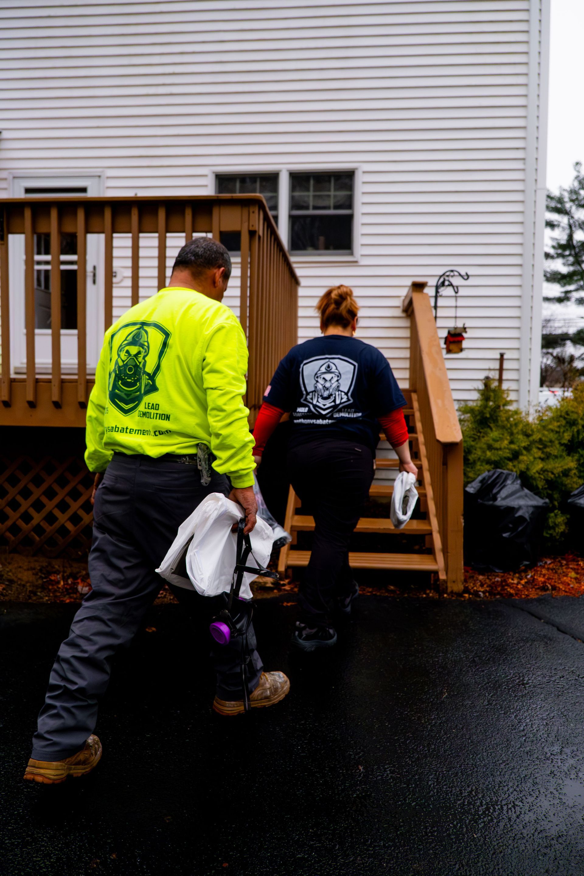 Two workers carrying items up wet steps beside a white house and wooden deck