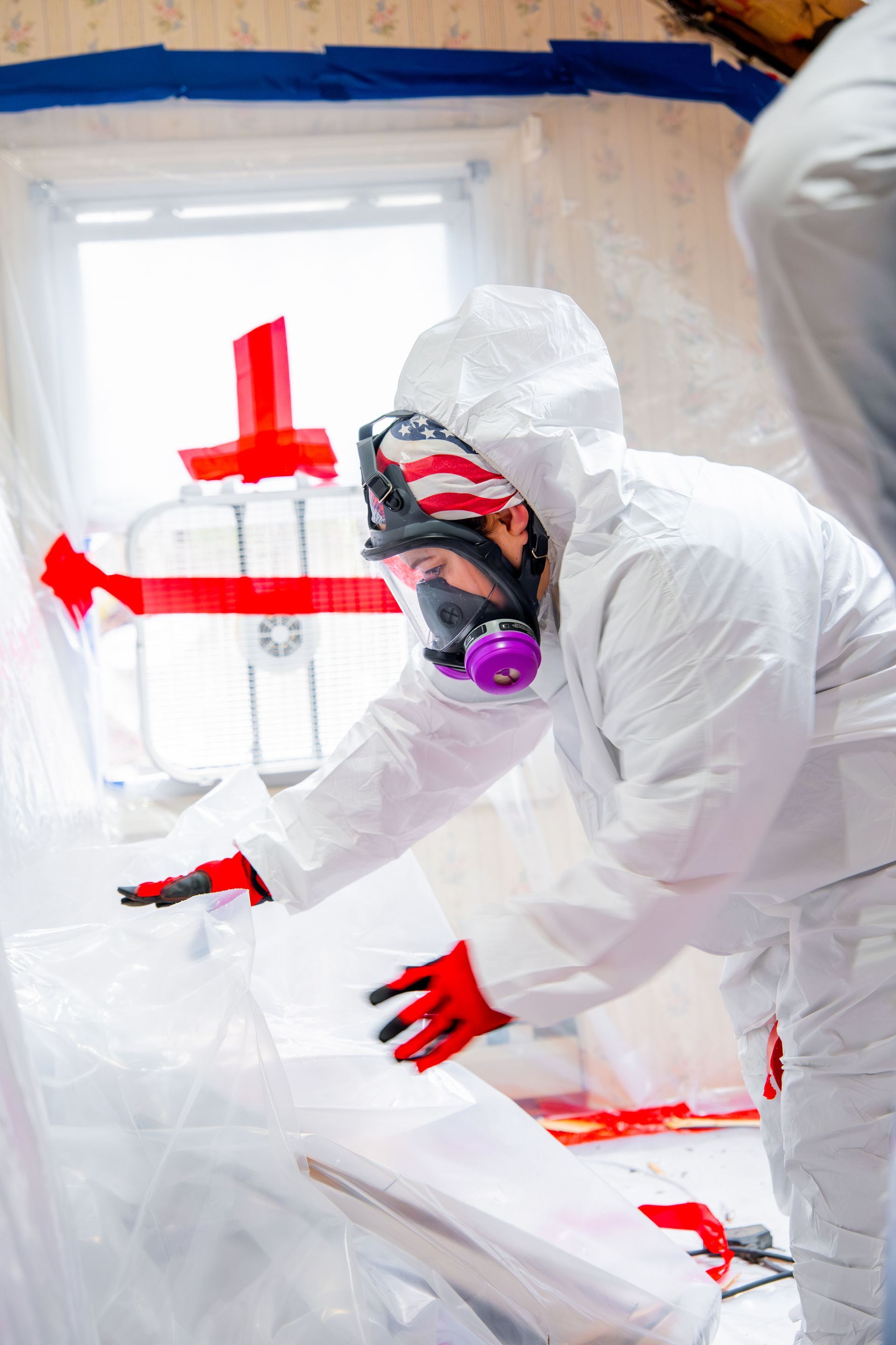 Workers in white hazmat suits painting a room, one wearing a respirator and red gloves