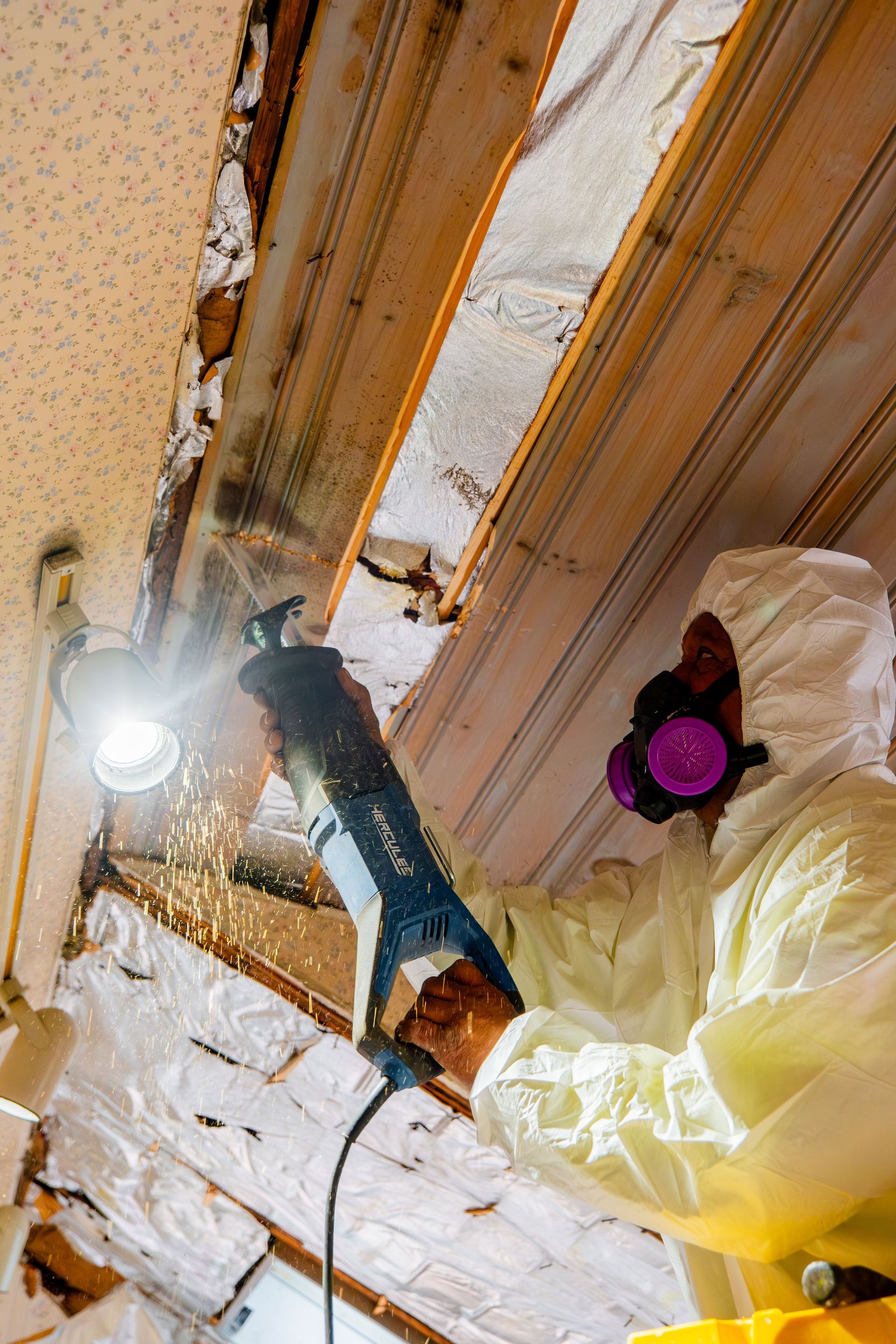Worker in protective gear using a power drill on a ceiling during renovation, with insulation exposed.