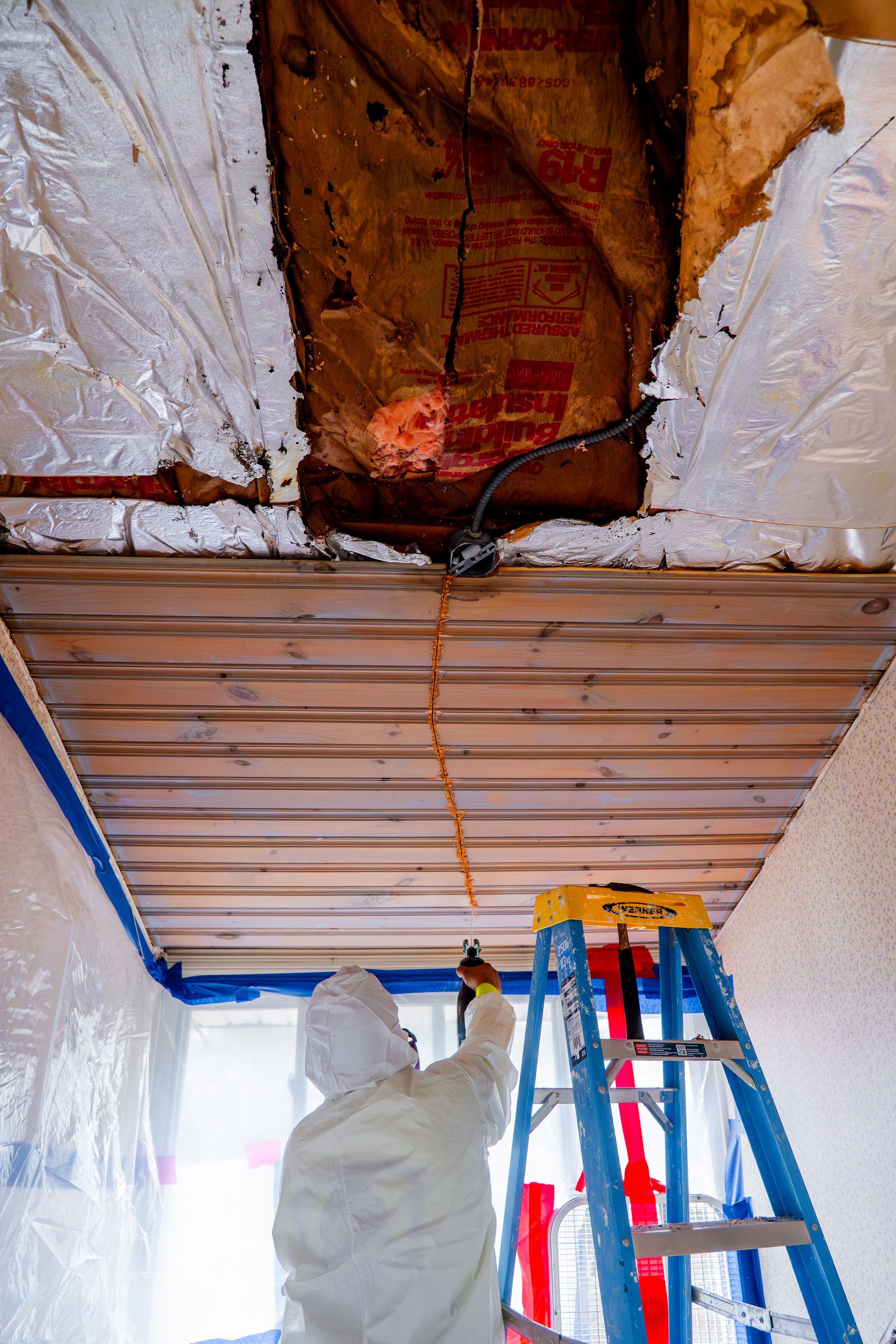 Worker using a ladder to repair a damaged ceiling with exposed brown insulation and hanging wires