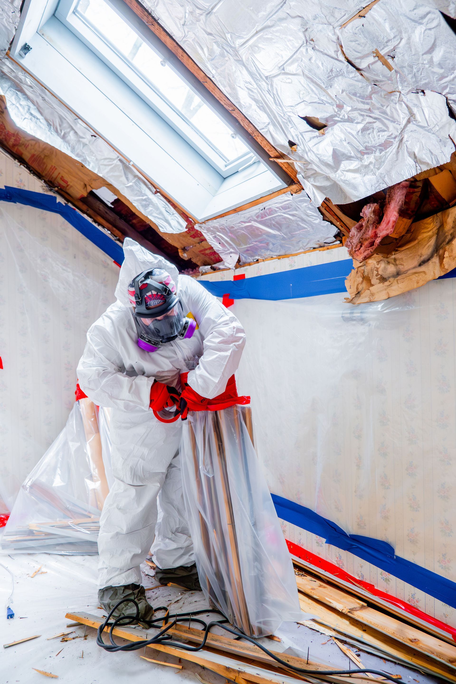 Worker in protective suit painting a room ceiling with plastic sheeting and blue tape masking the walls.