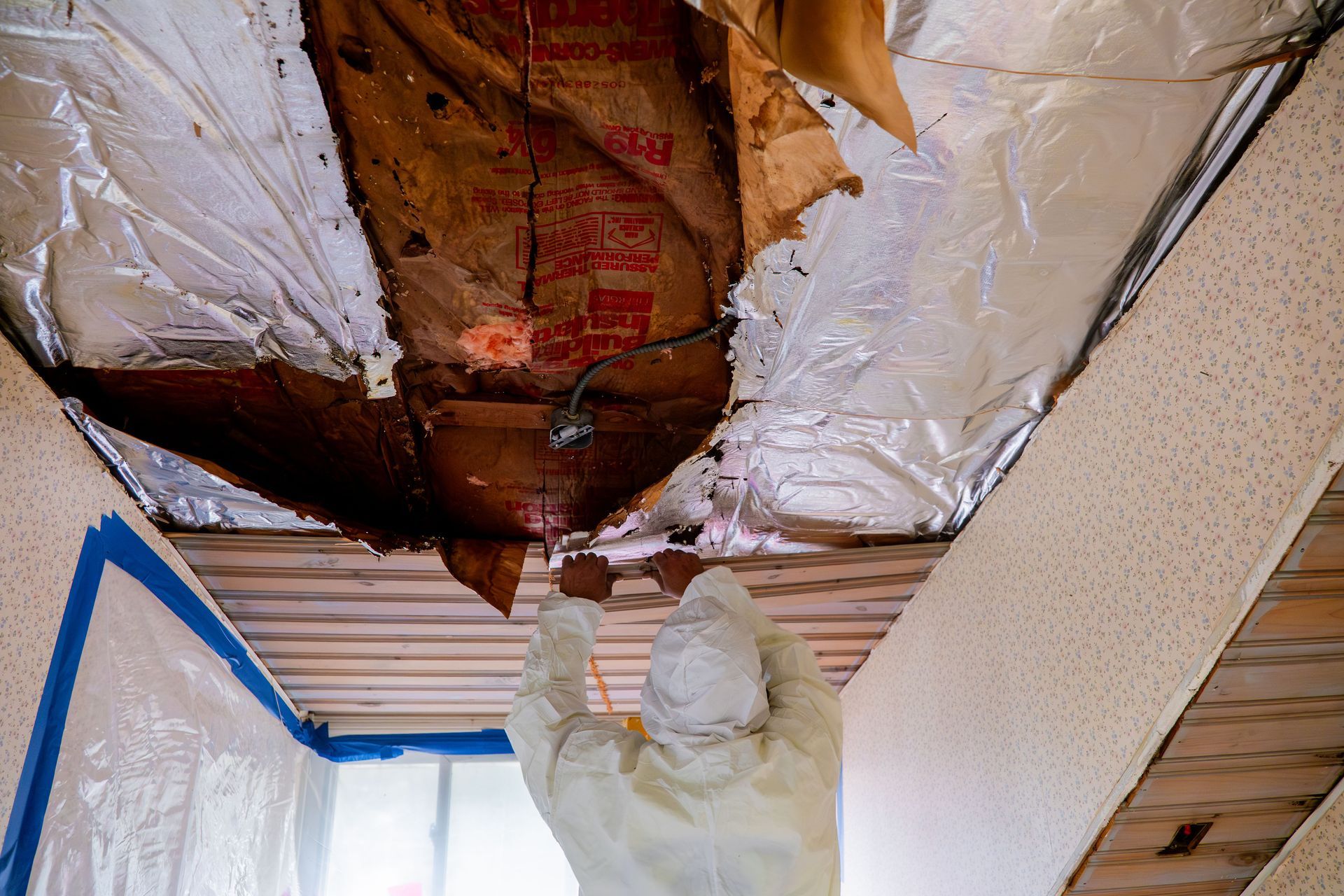 Person in protective suit removing damaged ceiling insulation during home repair