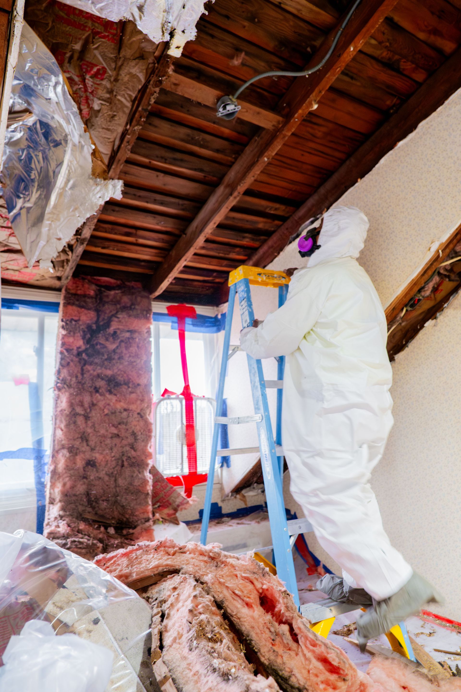 Worker in white protective suit removing insulation from a damaged ceiling inside a room