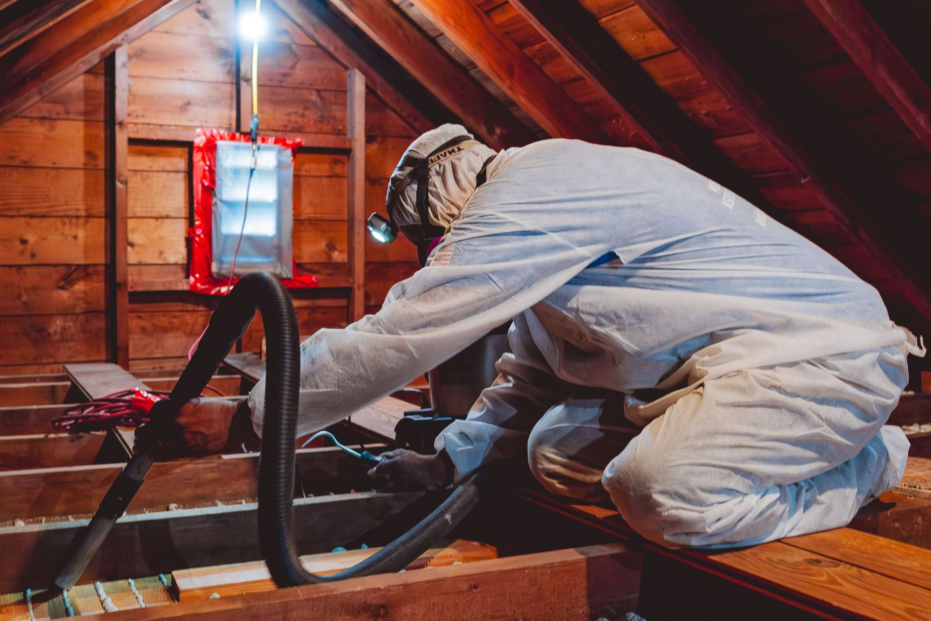 Worker in white coveralls inspecting attic insulation with a vacuum hose under a red-lit roof.