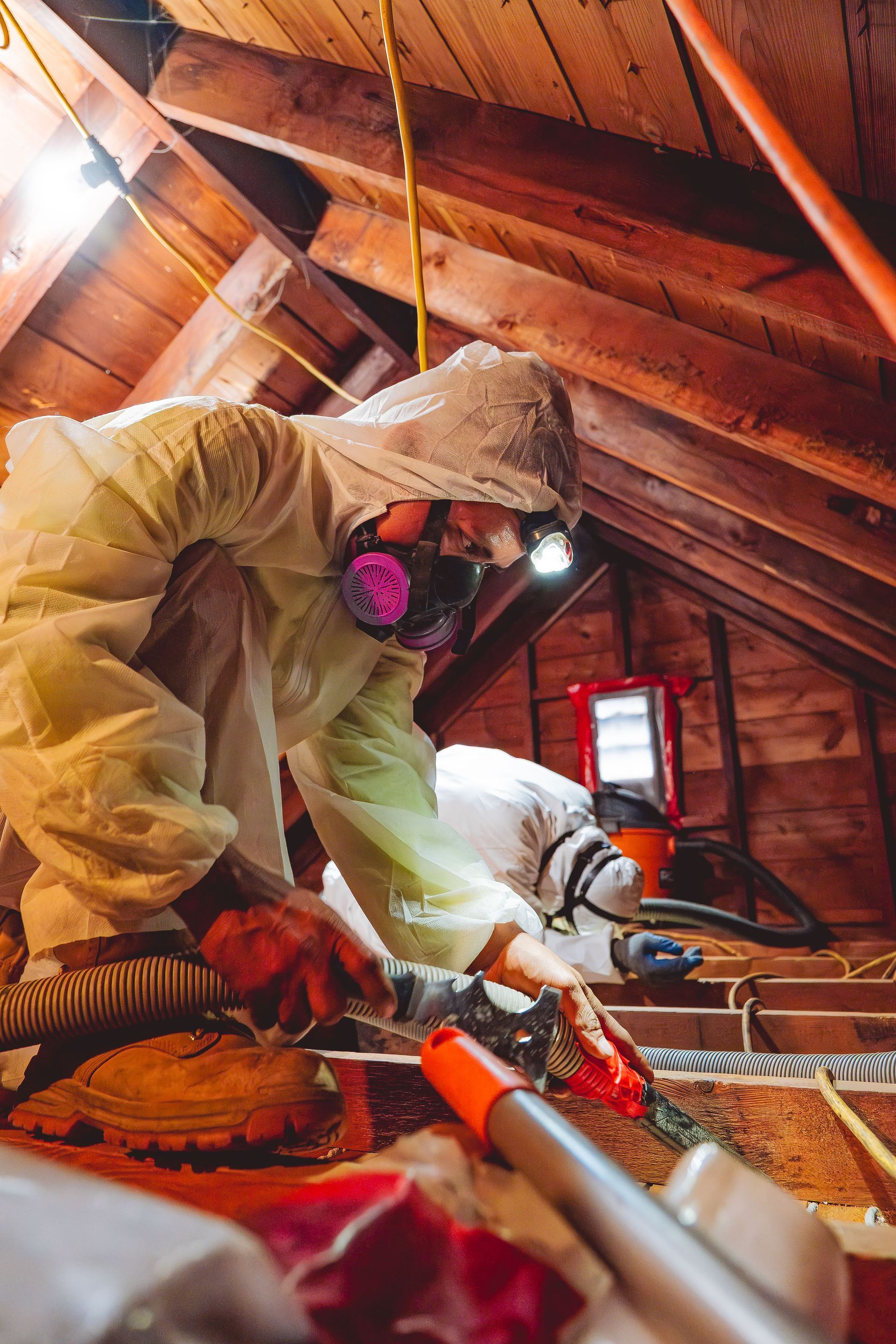 Two people in hazmat suits in an attic, removing insulation. One uses a tool, and both wear respirators.