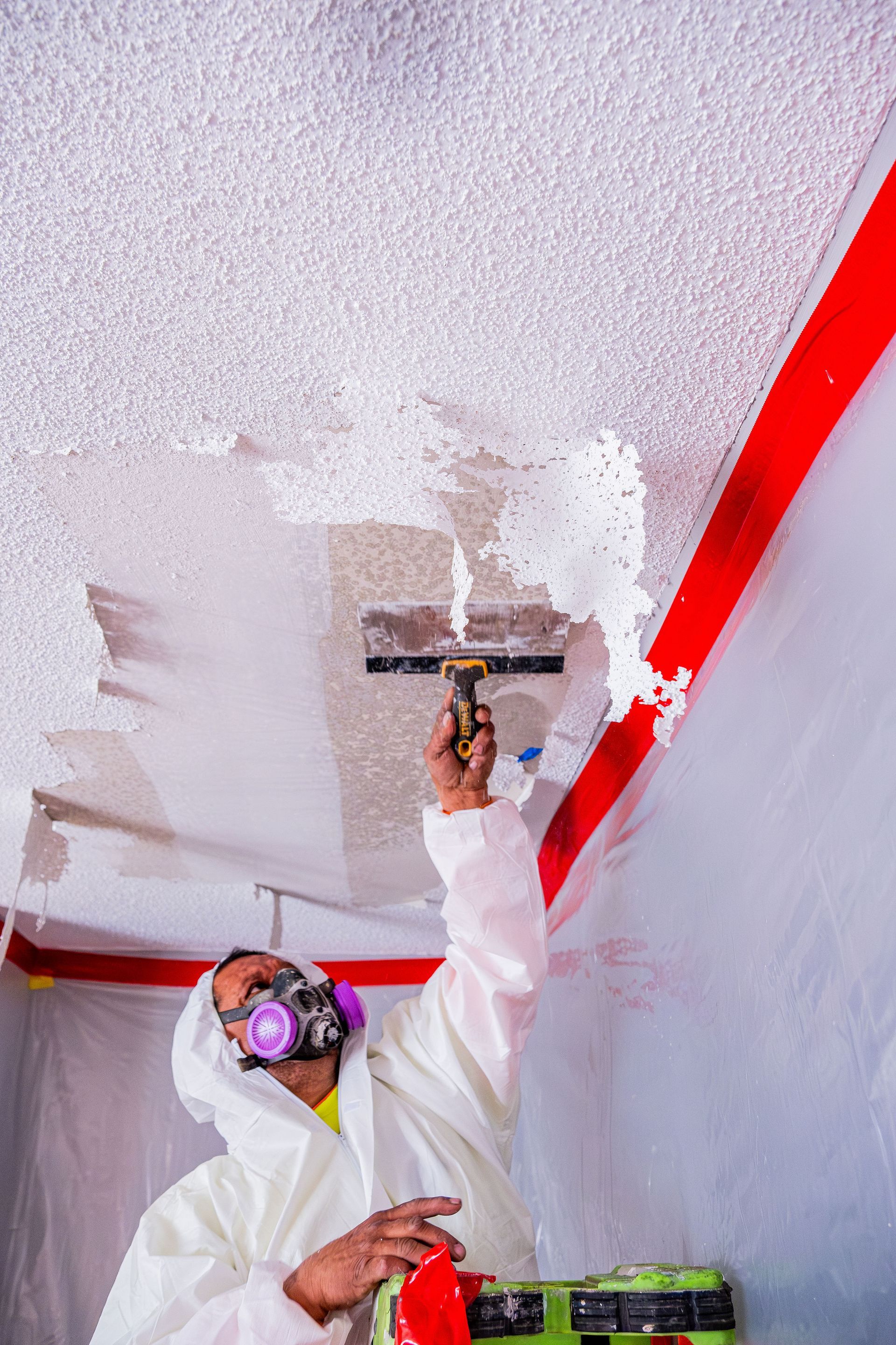 Person painting a ceiling with white paint using a roller, wearing a respirator and protective coveralls.