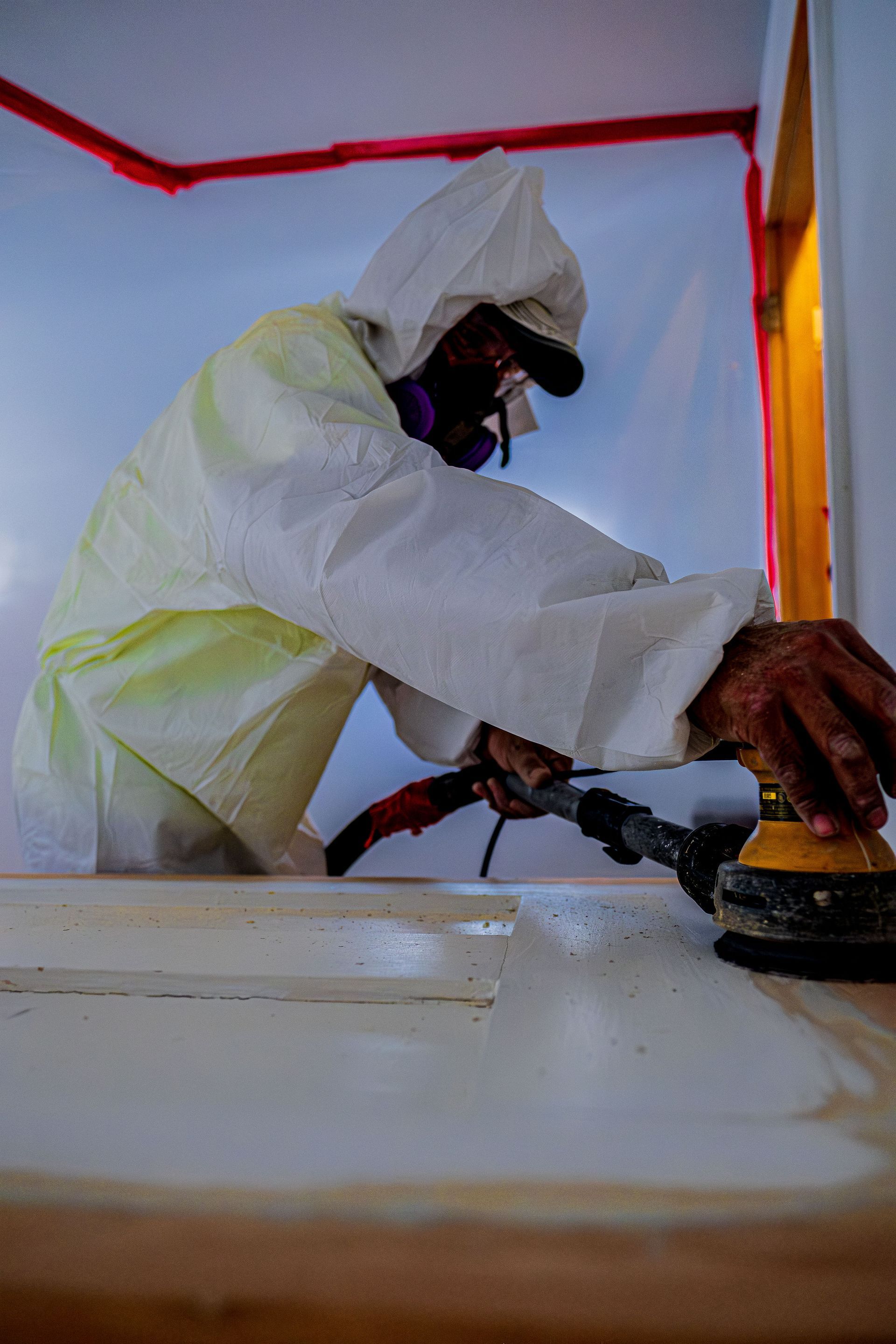 Worker in white protective suit sanding a wall inside a room with red trim and an open doorway