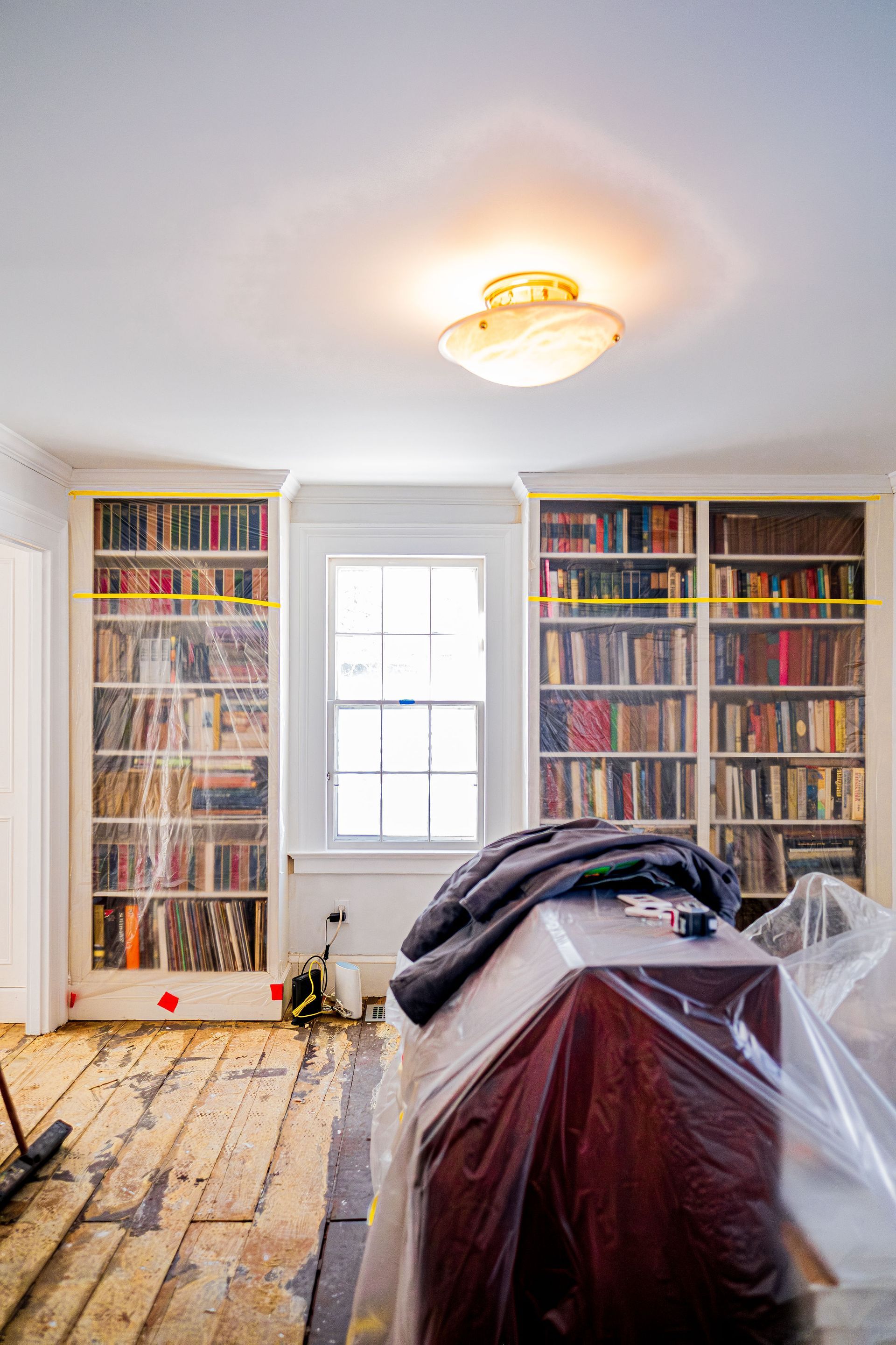 Bright room with tall bookshelves, a central window, and plastic-covered furniture during renovation.