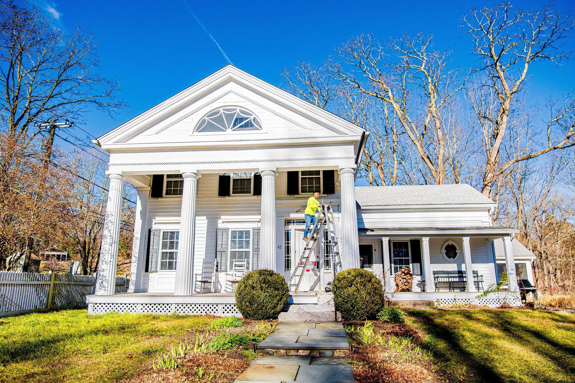 White colonial-style house with columns, blue shutters, and autumn trees under a clear sky