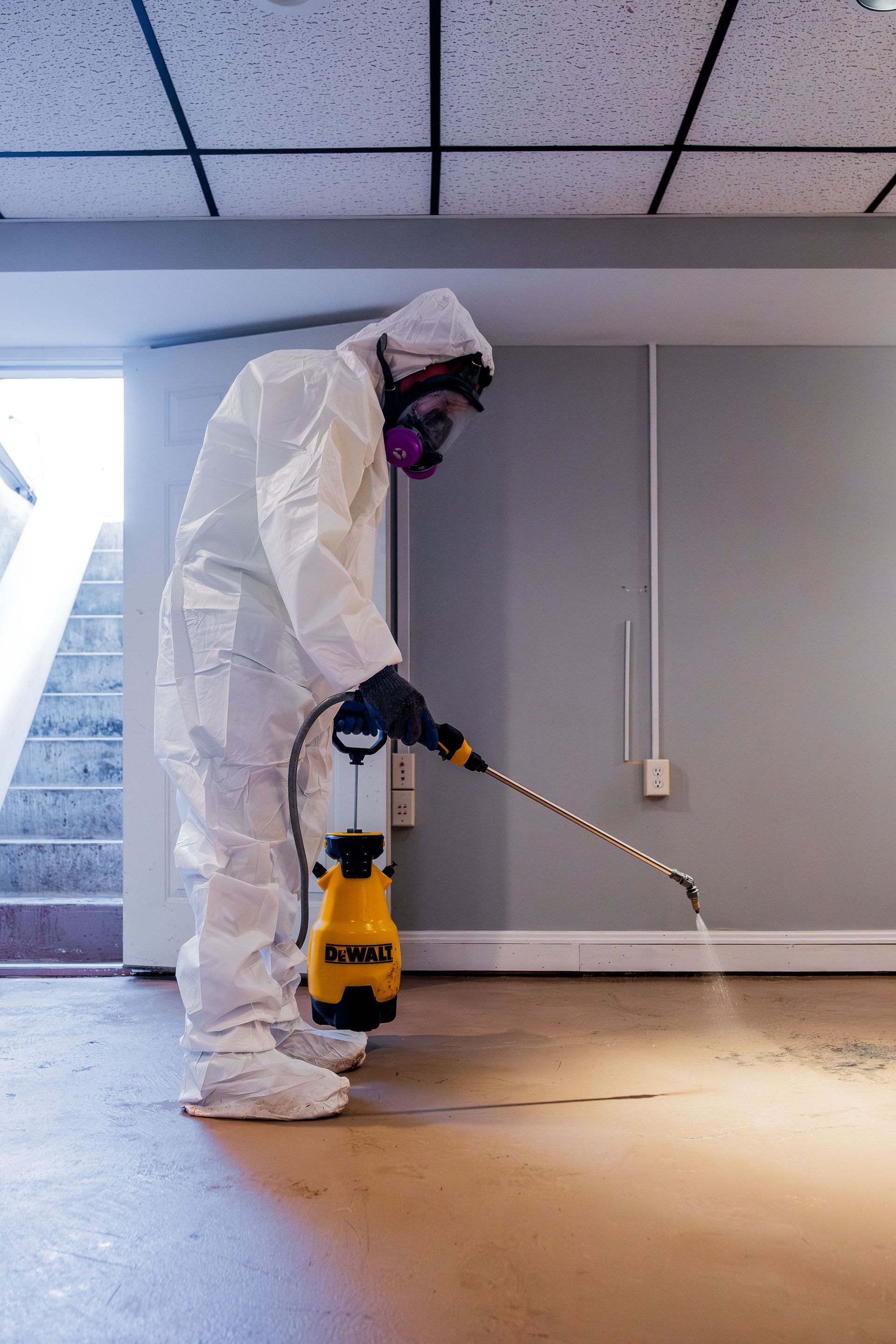 Person in protective suit spraying a floor with a yellow power sprayer indoors