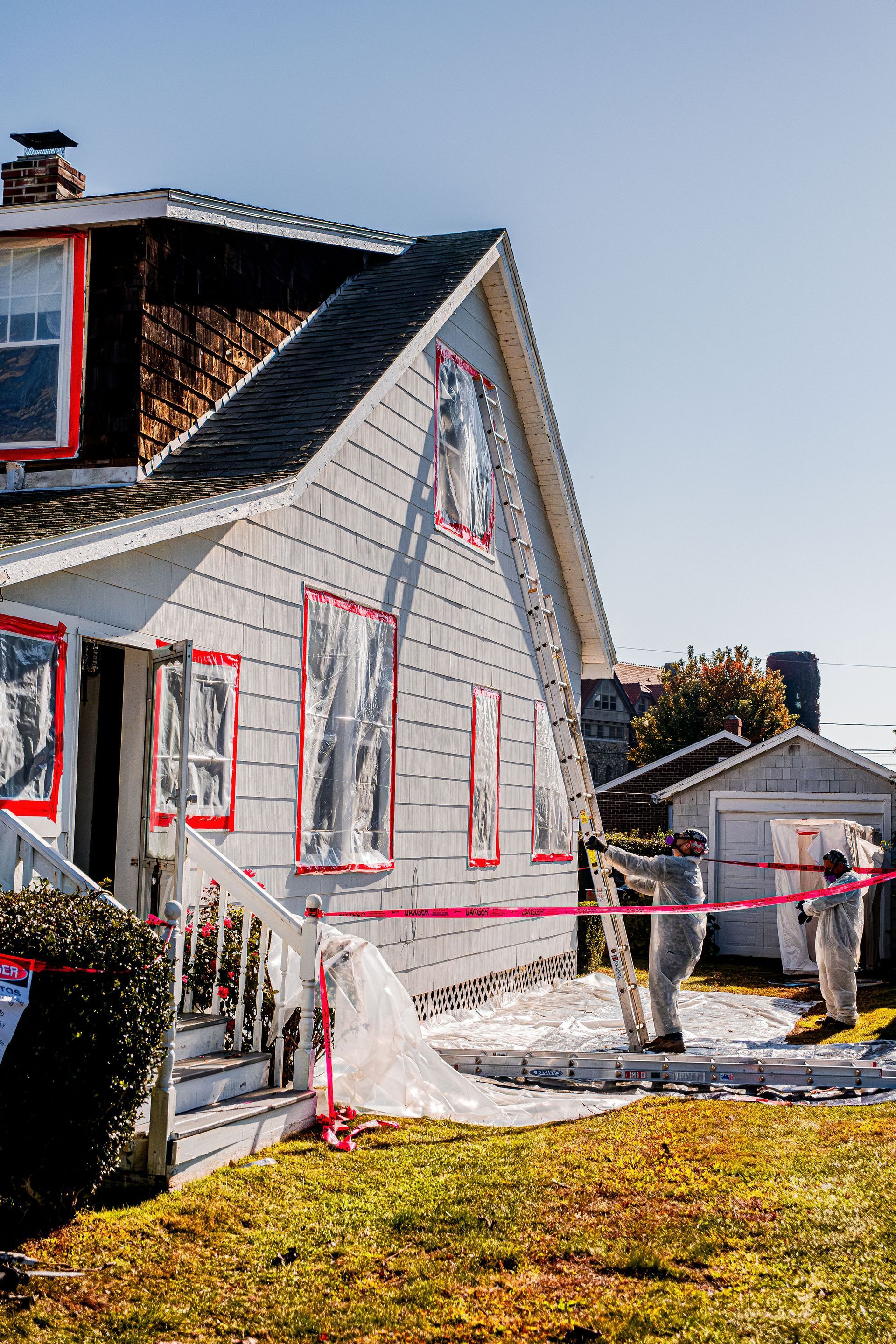 Damaged gray house with red fire tape and debris after a fire, with a charred roof and yard in front