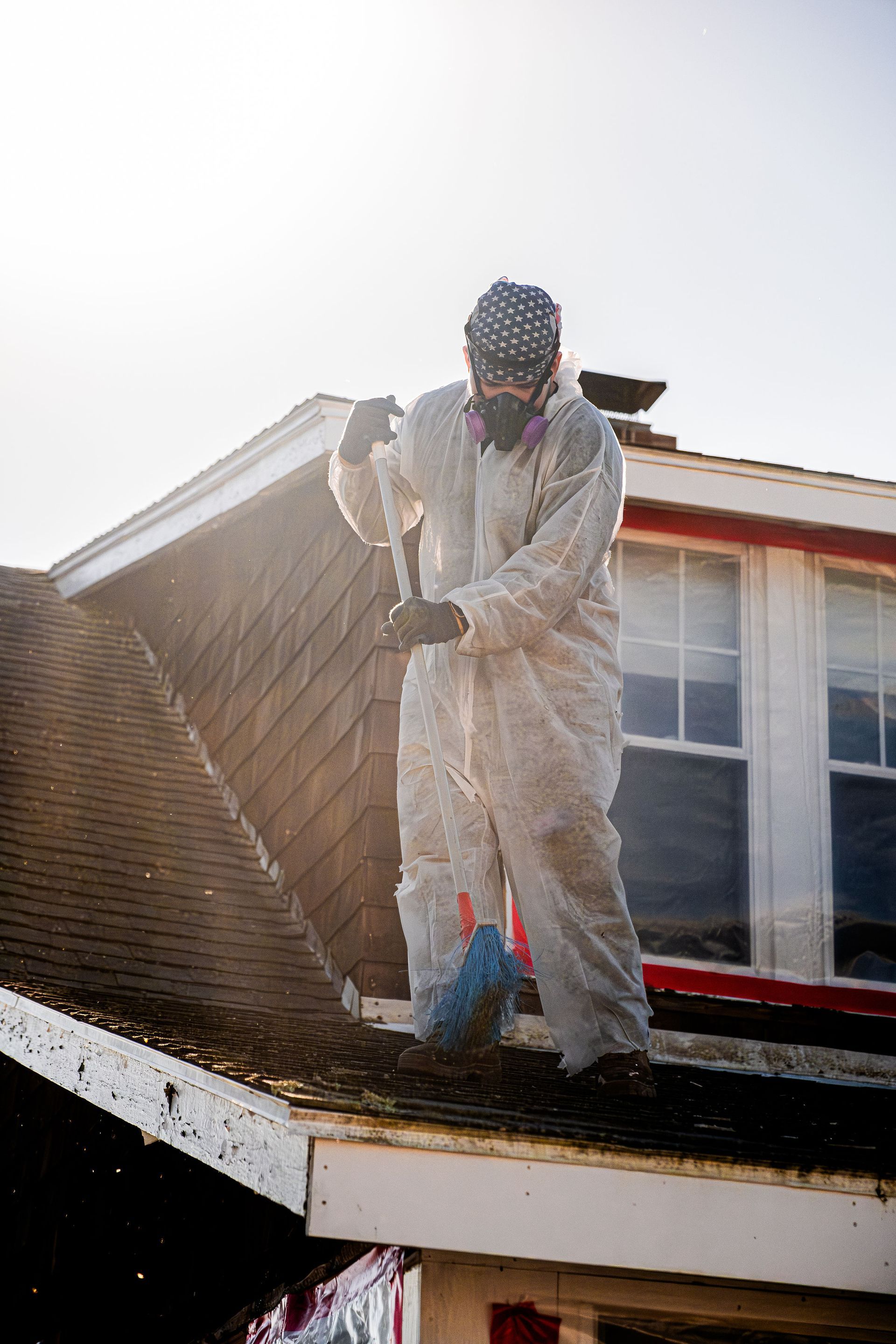 Person in protective suit power-washing a roof beside a house window.