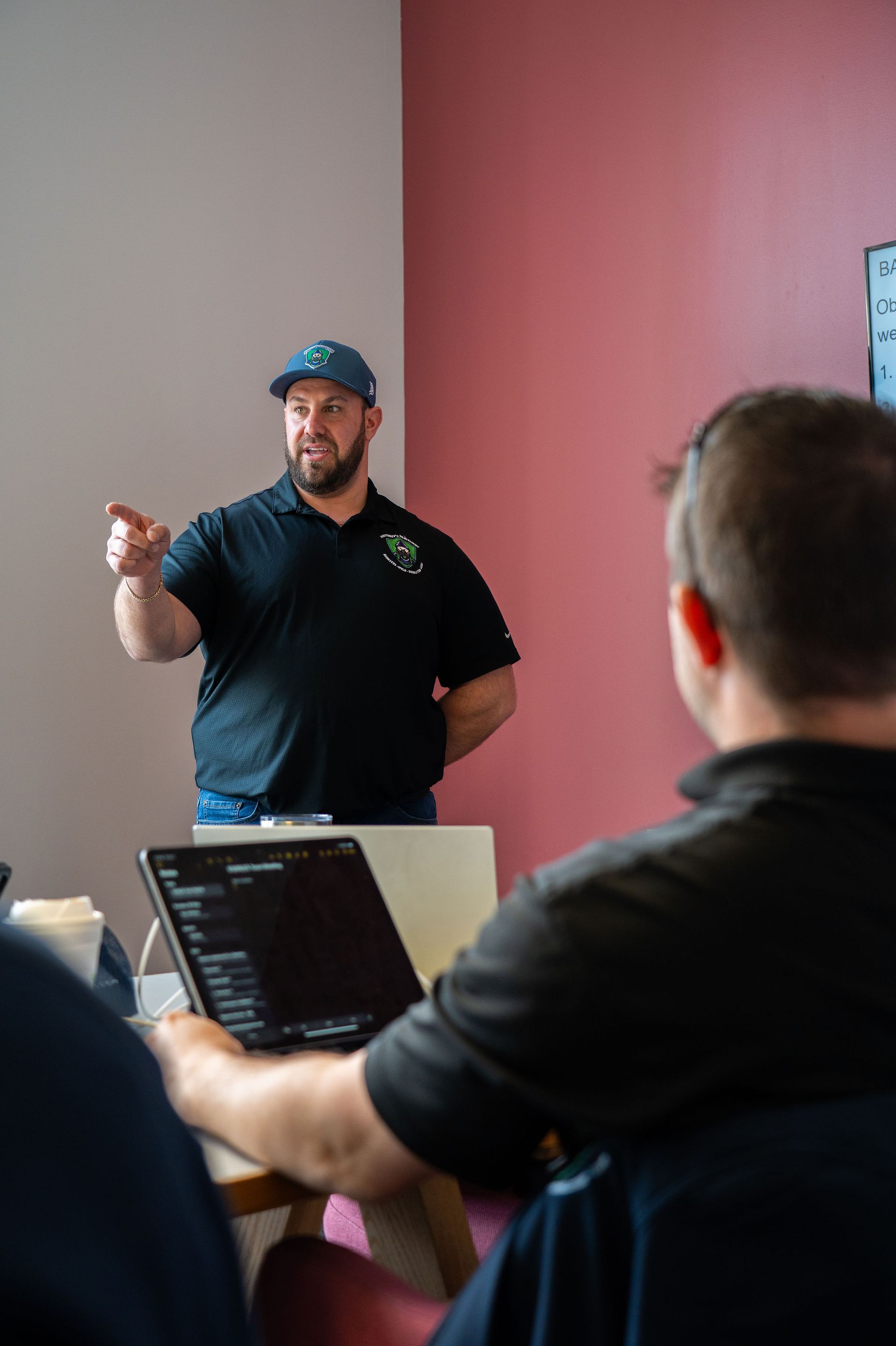 Man speaking to a small audience in a meeting room, pointing toward a laptop on a table.