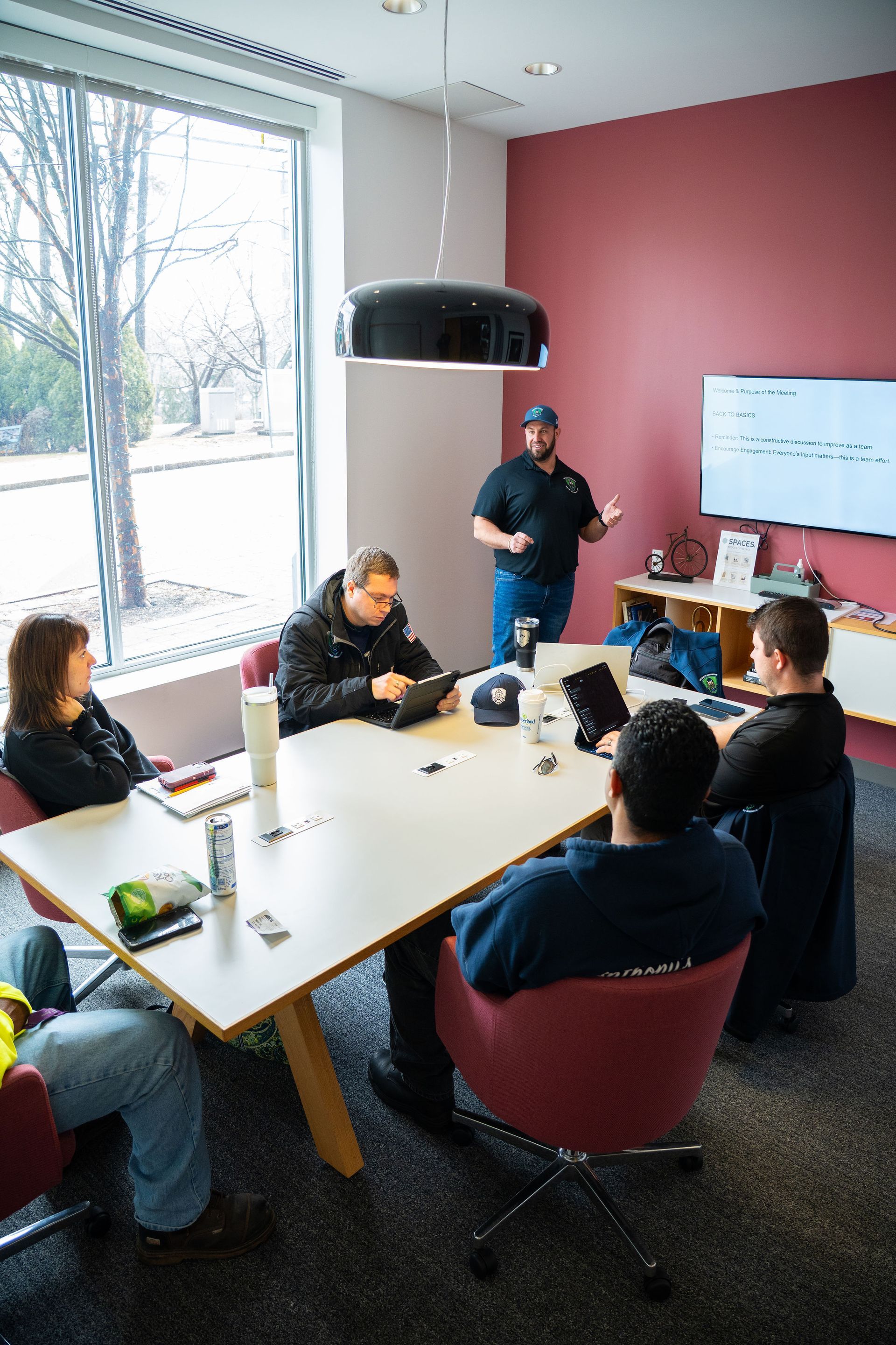 Small meeting room with five people around a table and one person presenting beside a whiteboard