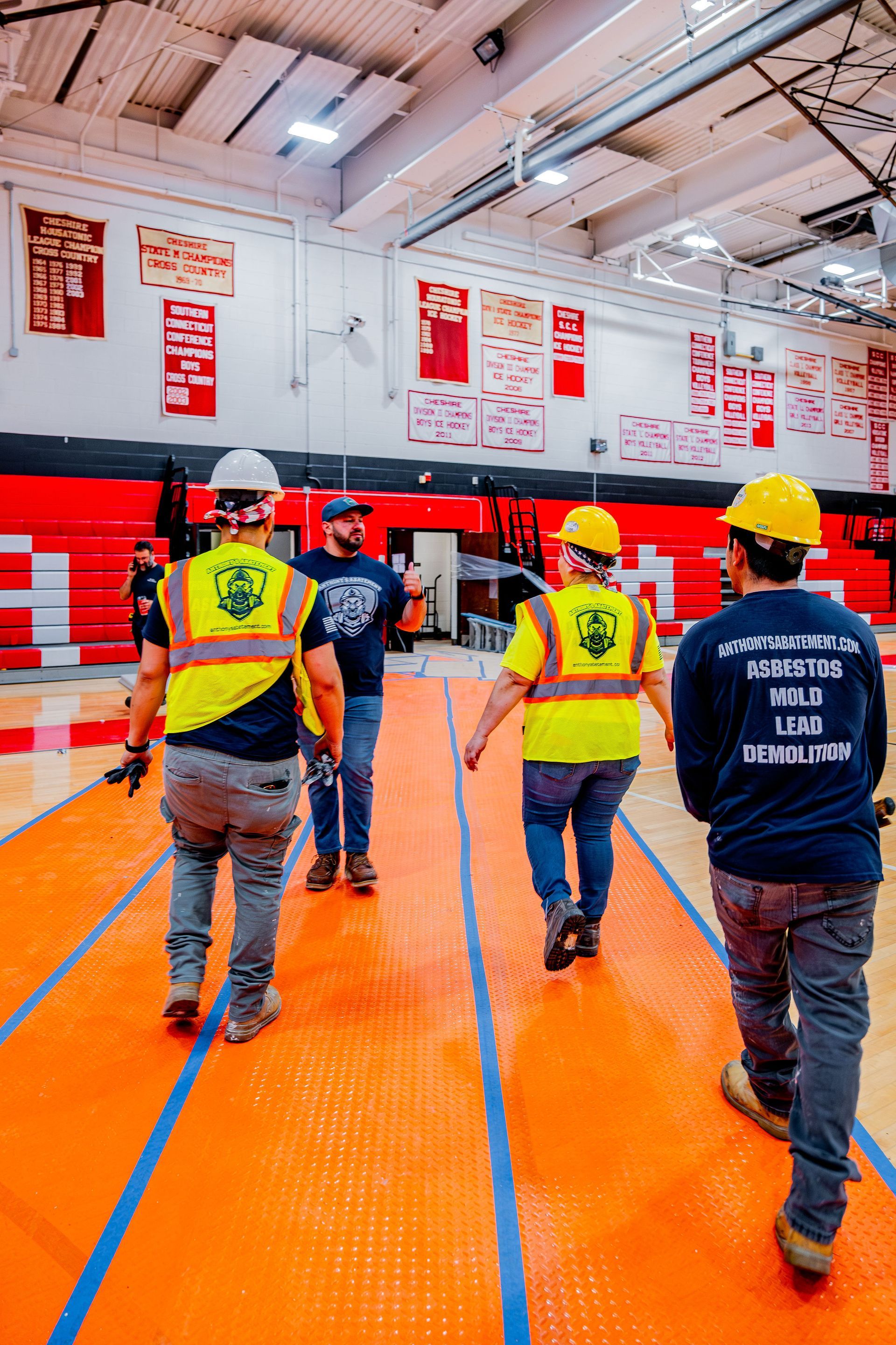 Workers in yellow helmets walk across an orange gym floor with red bleachers in the background