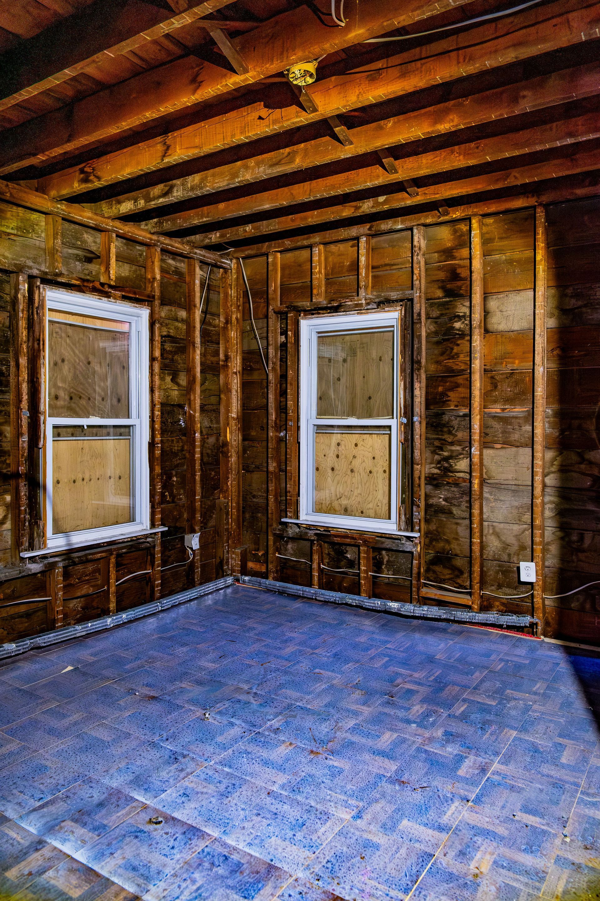 Empty unfinished room with exposed wooden walls, ceiling beams, two windows, and a blue floor covering