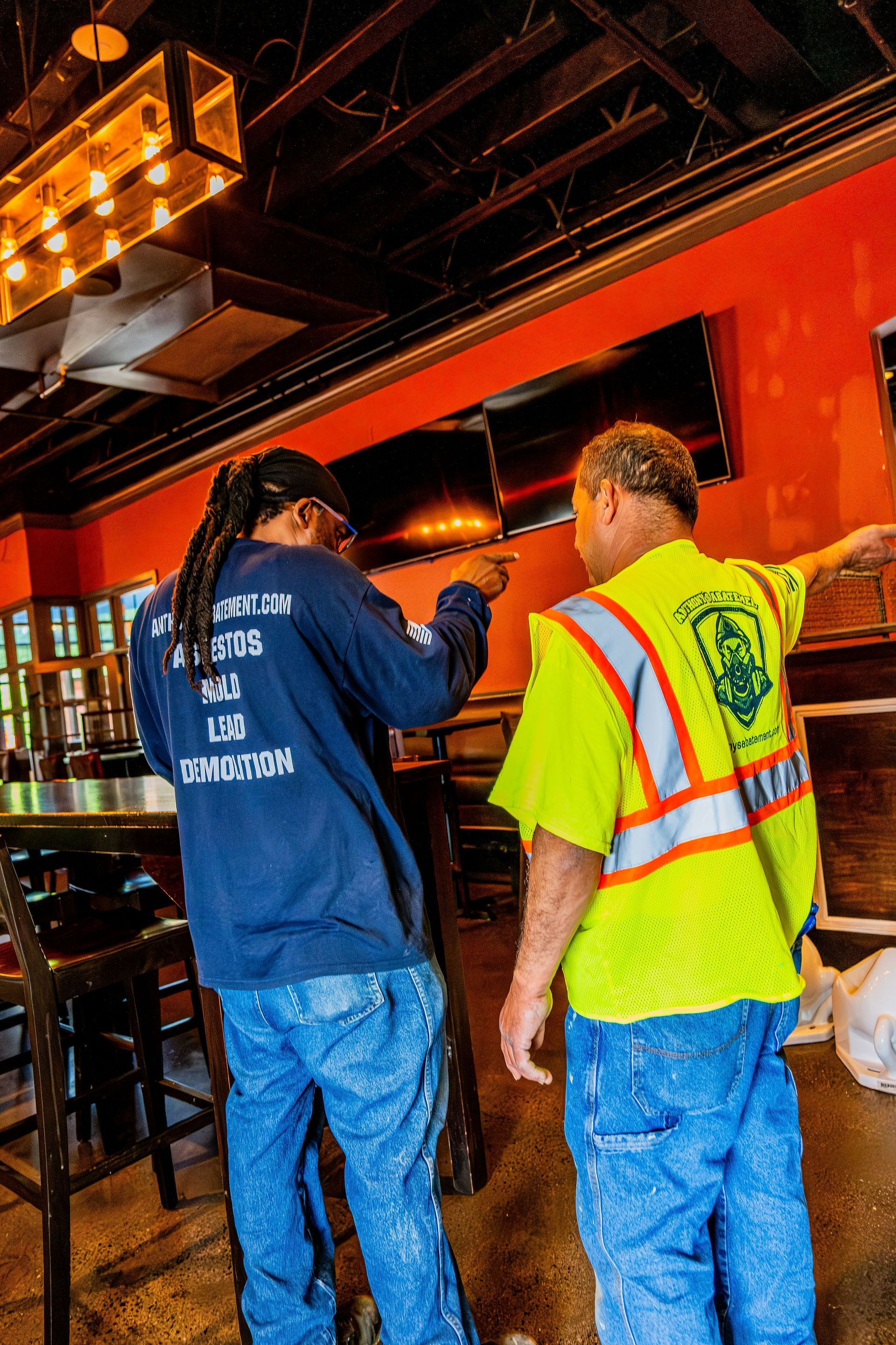 Two workers in a bar interior, one in a blue shirt and one in a neon safety vest, talking near the counter