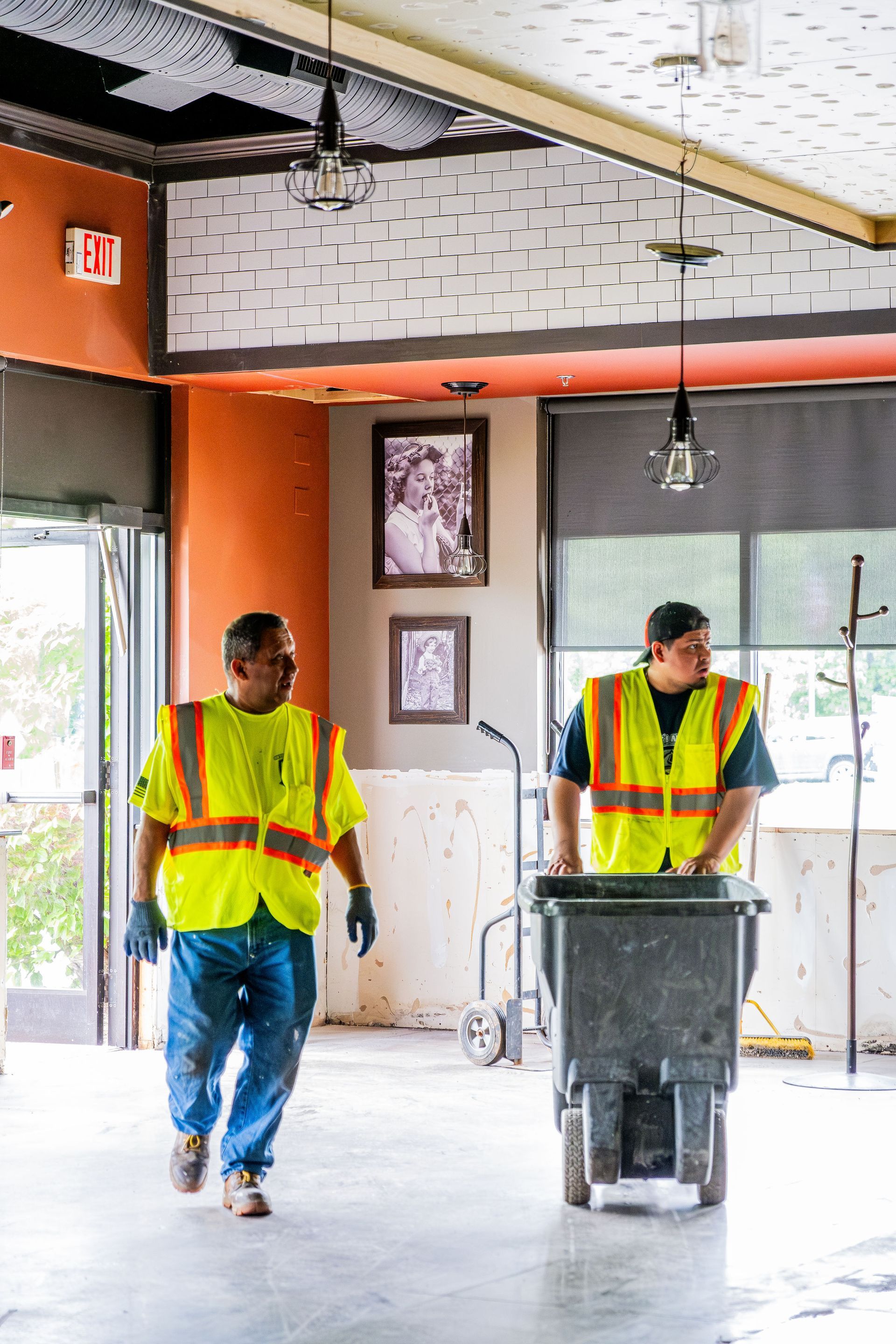Two construction workers in high-vis vests move a trash bin through an indoor renovation site