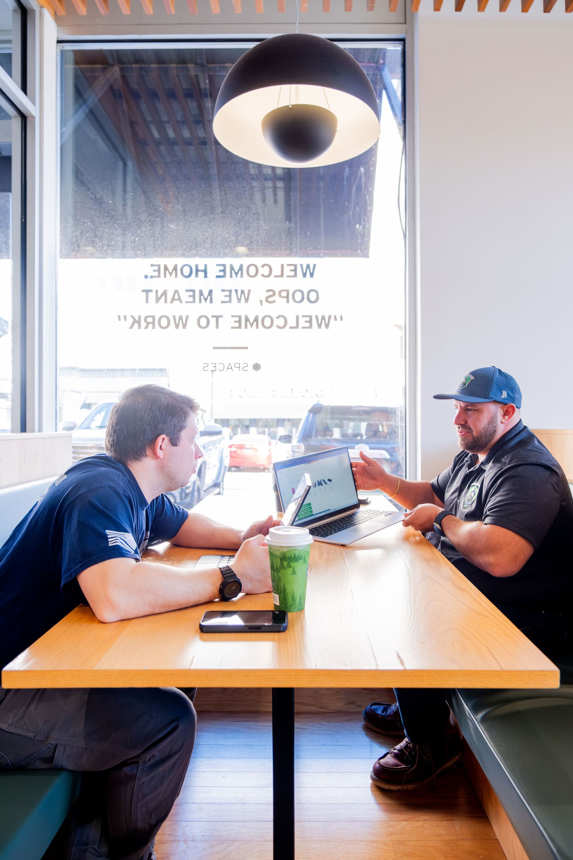 Two men talking over laptops at a café table with a large window and hanging light above them
