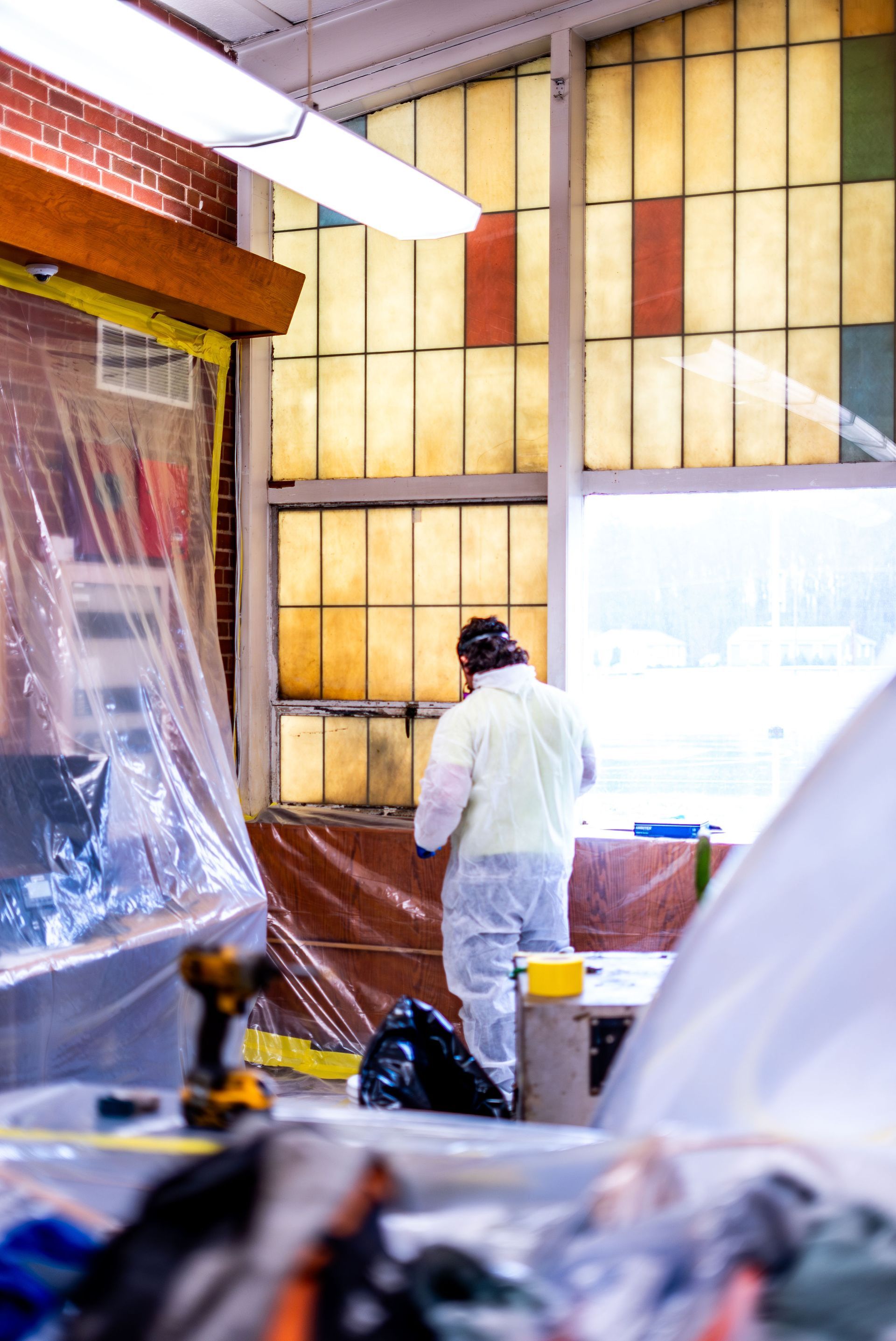 Worker in white coveralls stands in a room under renovation with plastic-covered furniture and bright windows.