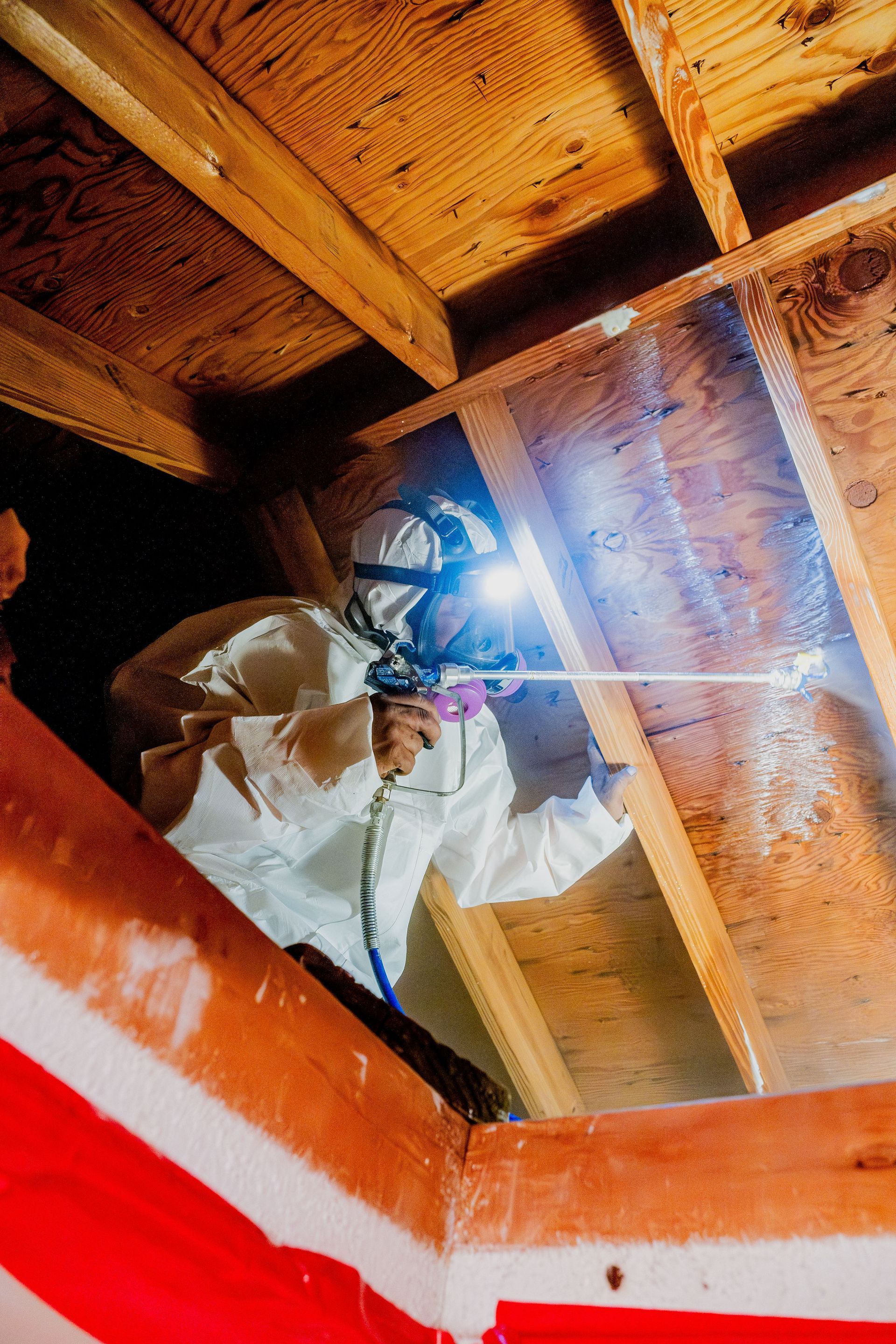 Worker in protective gear spraying foam insulation inside a wooden attic.