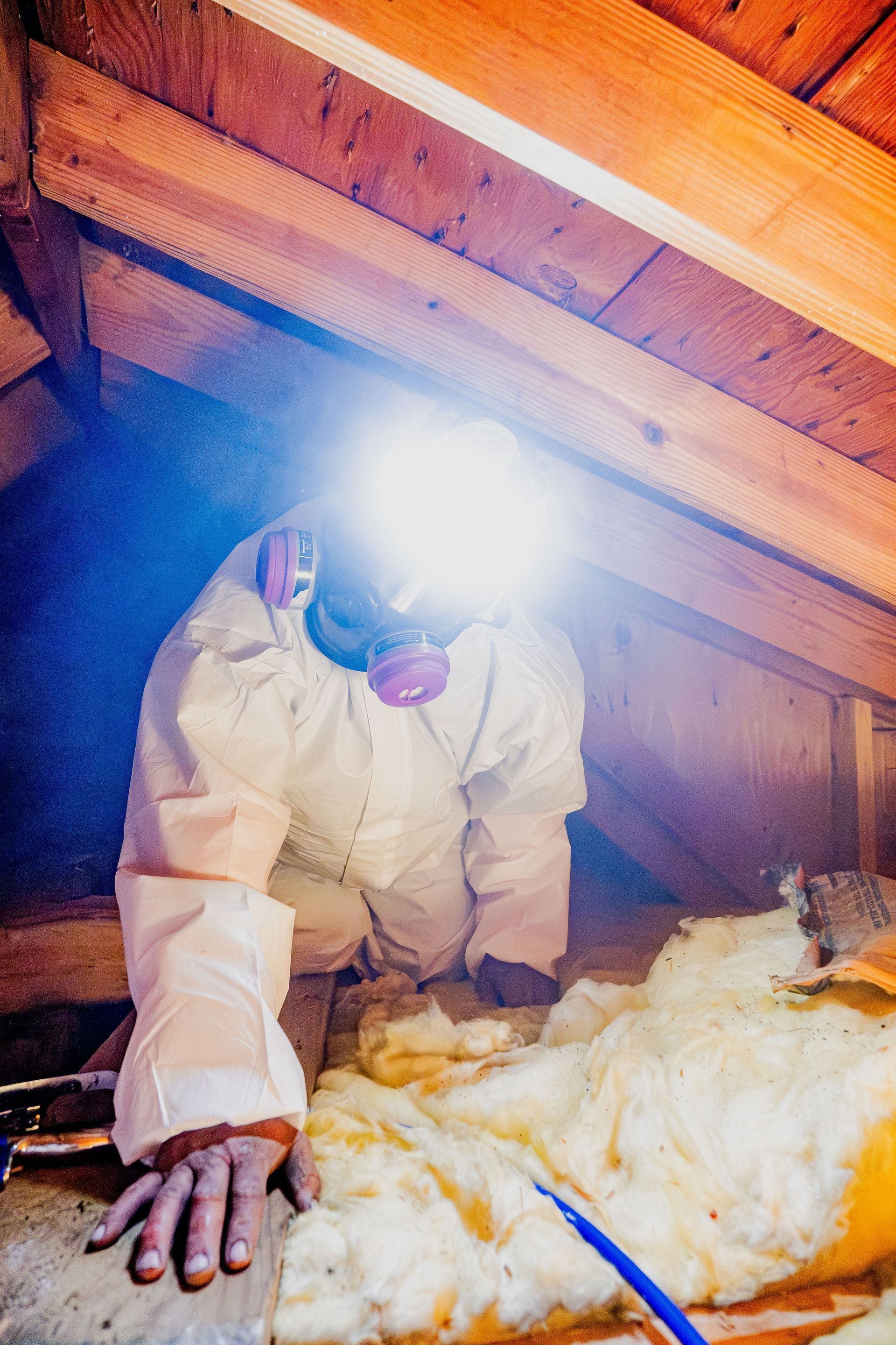 Person in protective suit inspecting attic insulation with a bright headlamp.