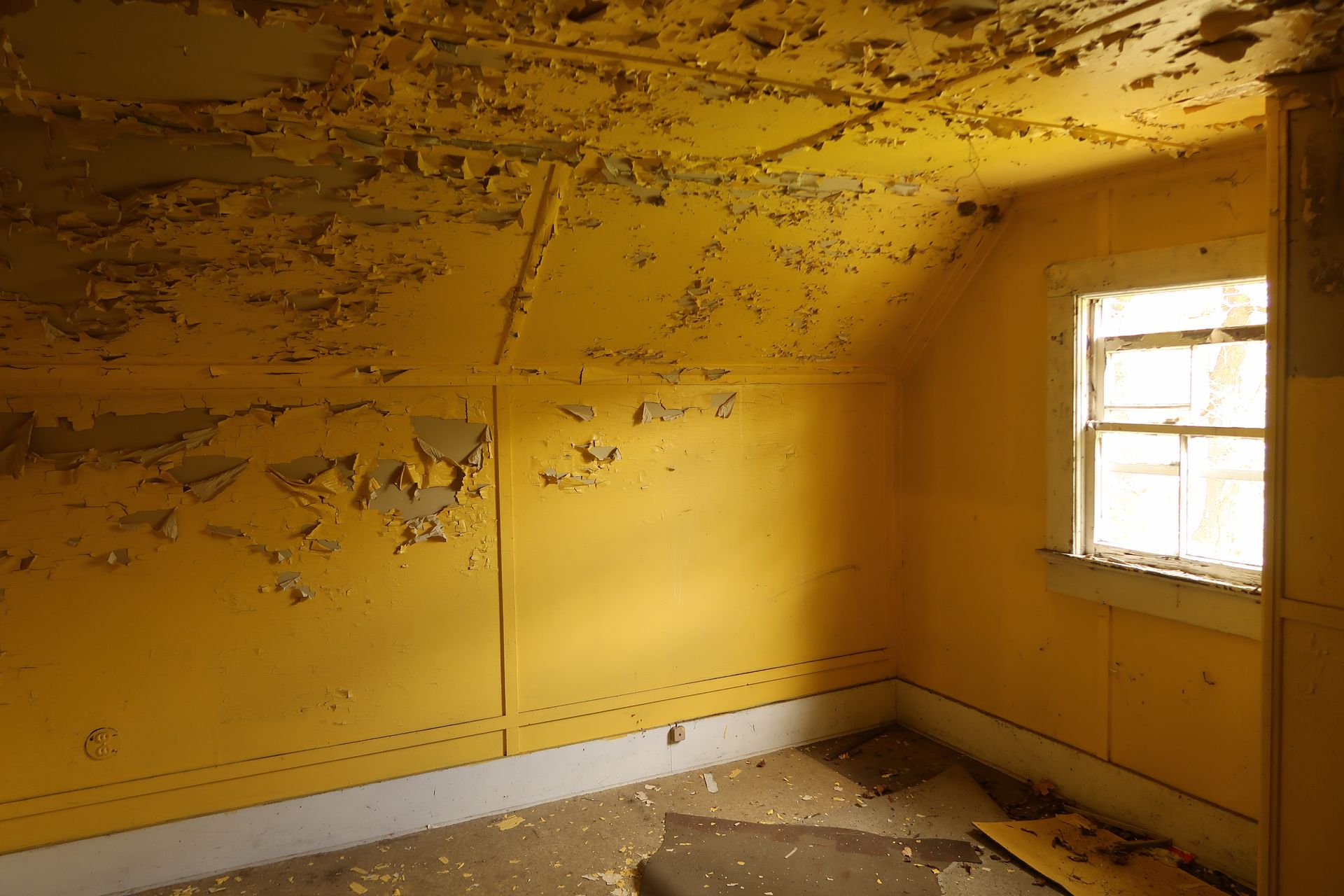 Peeling yellow room with damaged walls, ceiling, and a window letting in light