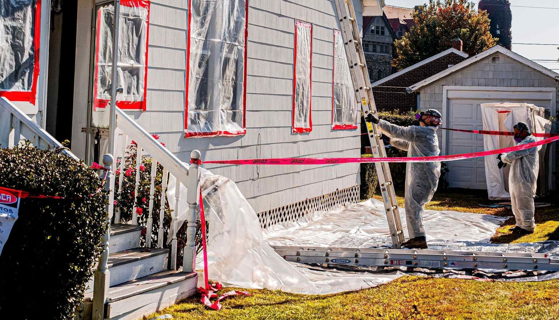 House exterior wrapped in white plastic and red tape, with a worker near a small shed and ladder.