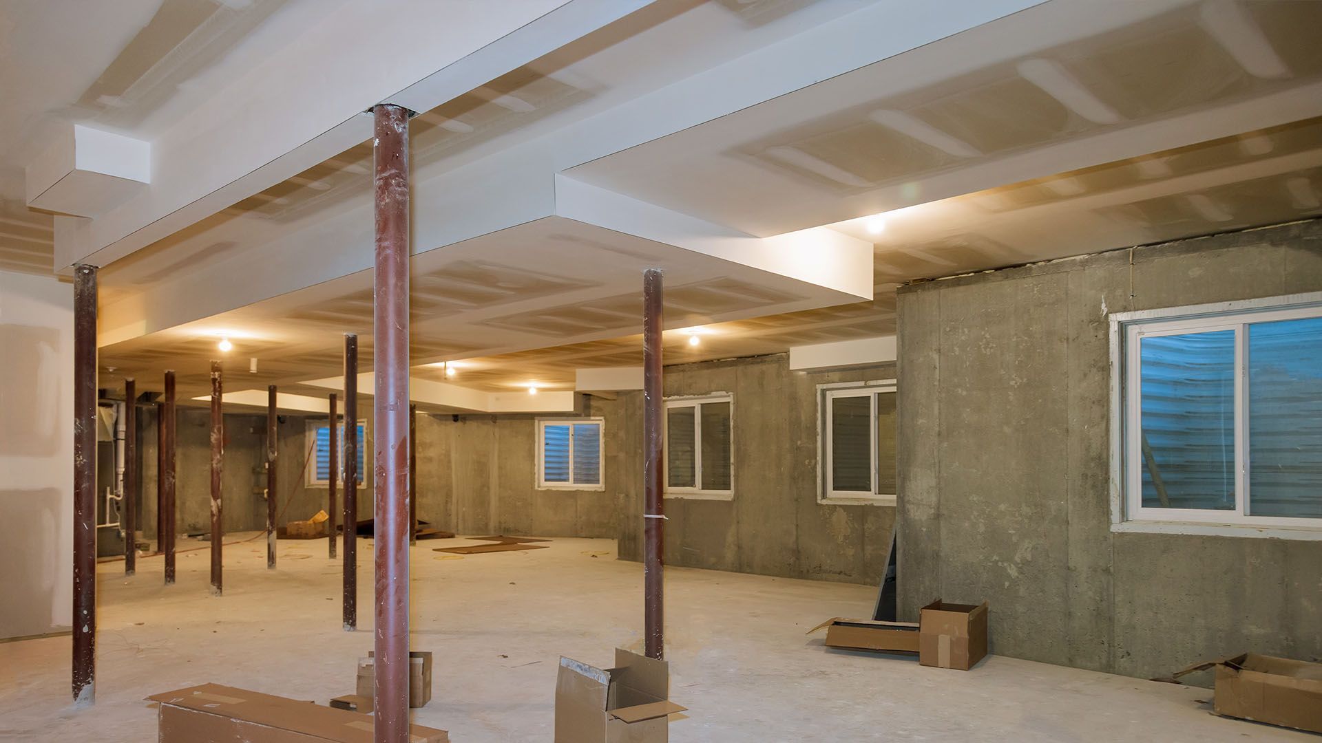 Basement under renovation with support beams, drywall ceiling, concrete walls, and windows.