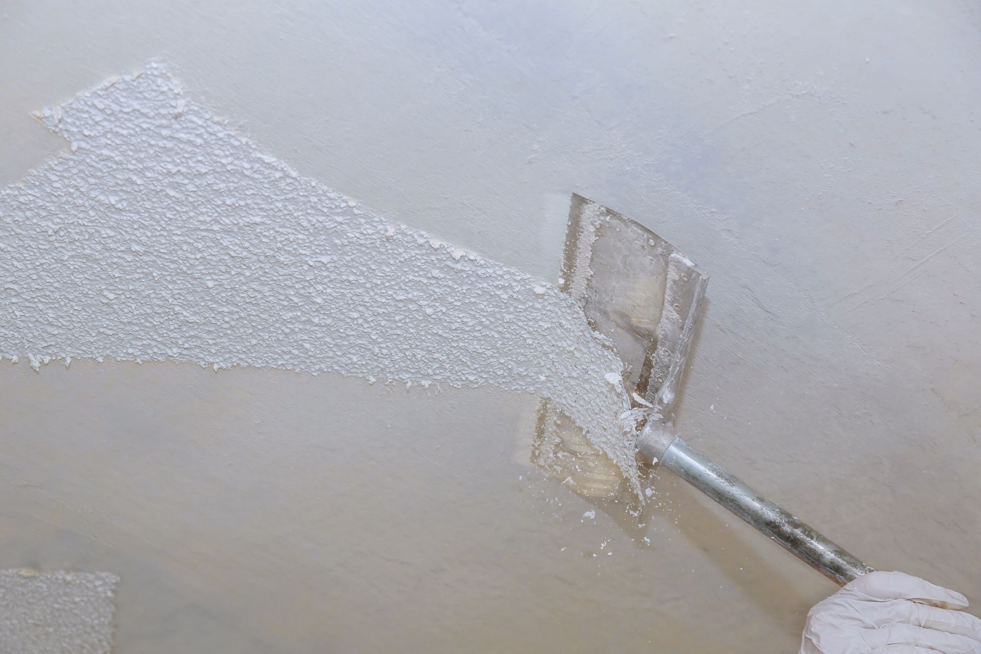 A metal scraper removes textured white popcorn finish from a wall, revealing the smooth surface underneath.