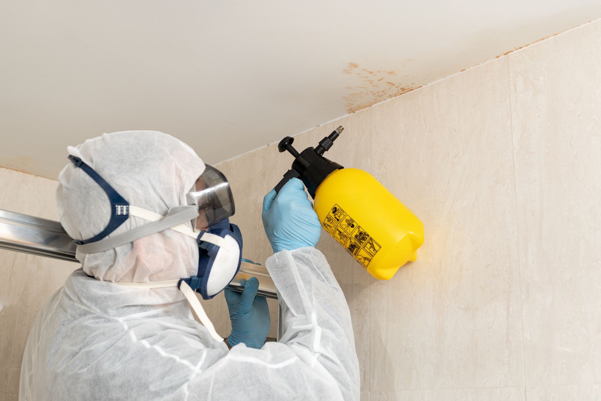 Worker in protective suit sprays pesticide on a wall with a yellow sprayer and blue gloves.