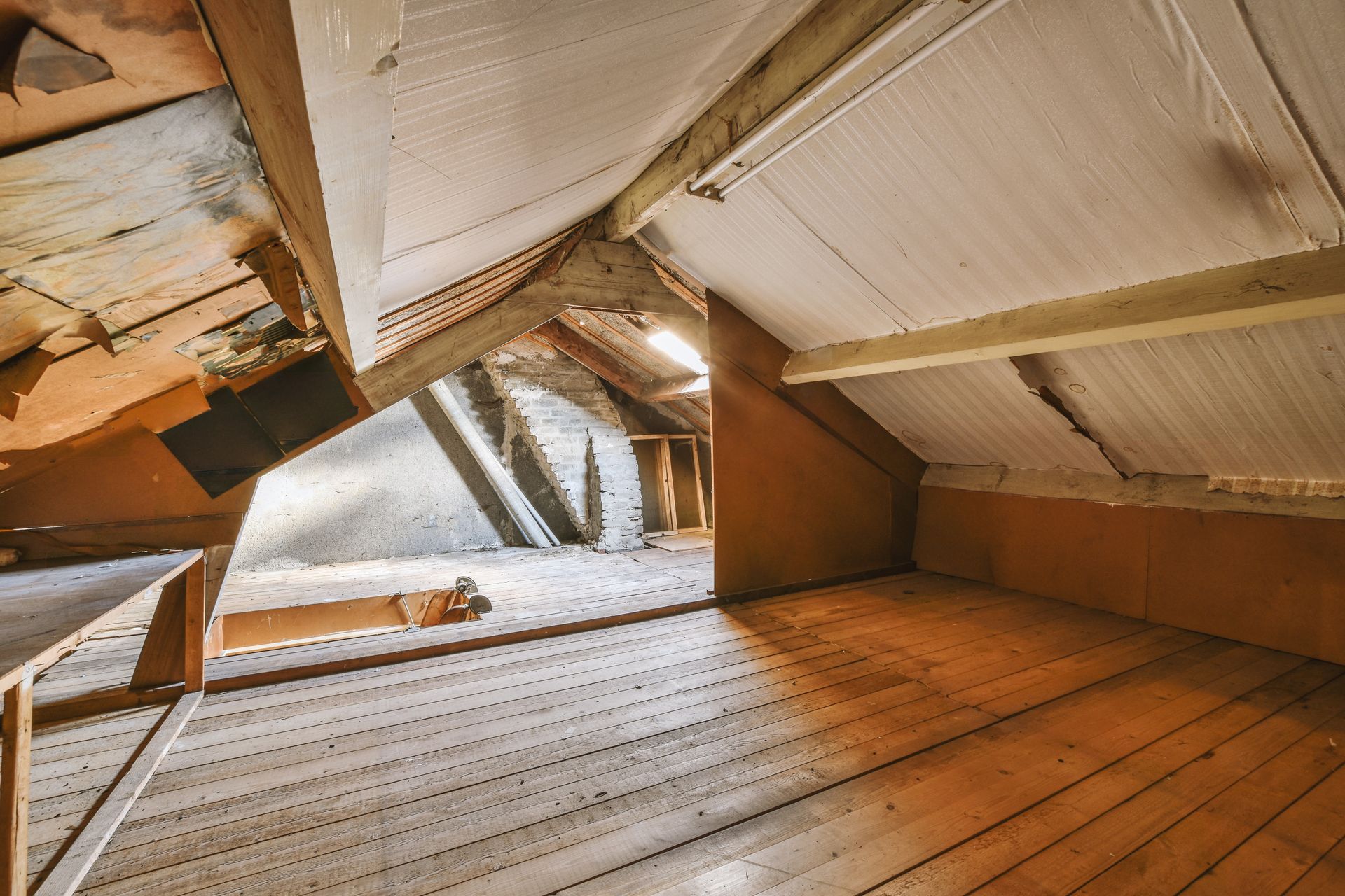 Abandoned attic with slanted wooden beams, peeling ceiling, and sunlight through a small opening