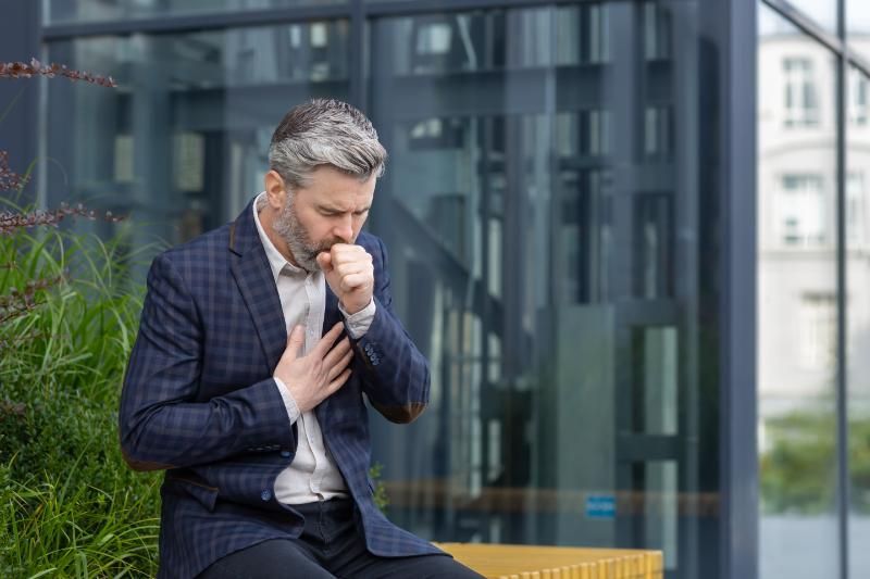 A man in a suit is sitting on a bench and coughing.