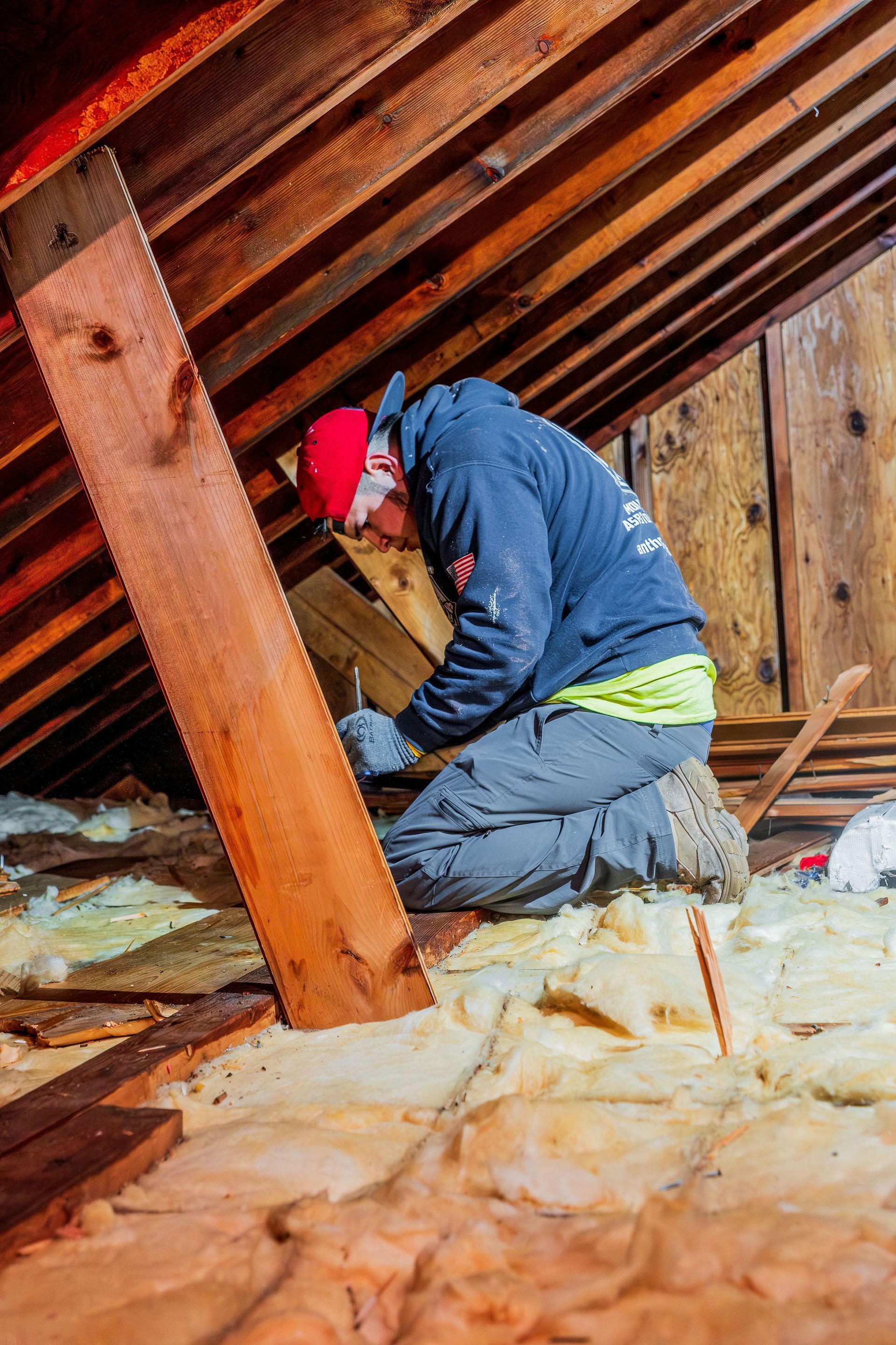 Worker kneeling in an attic insulation space, wearing a red hard hat and blue jacket.