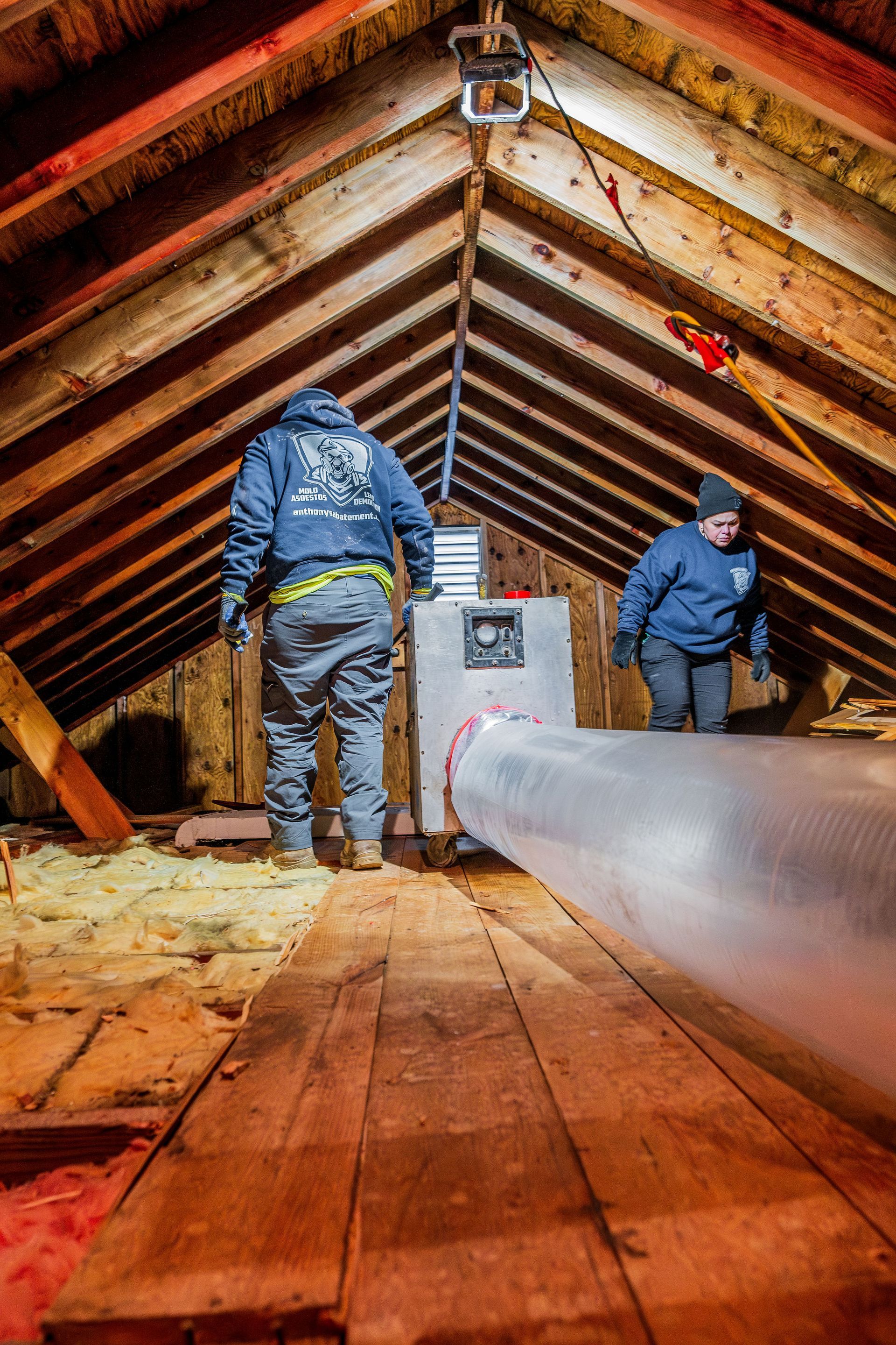 Two workers in a wooden attic inspect a large gray duct beneath the roof beams.