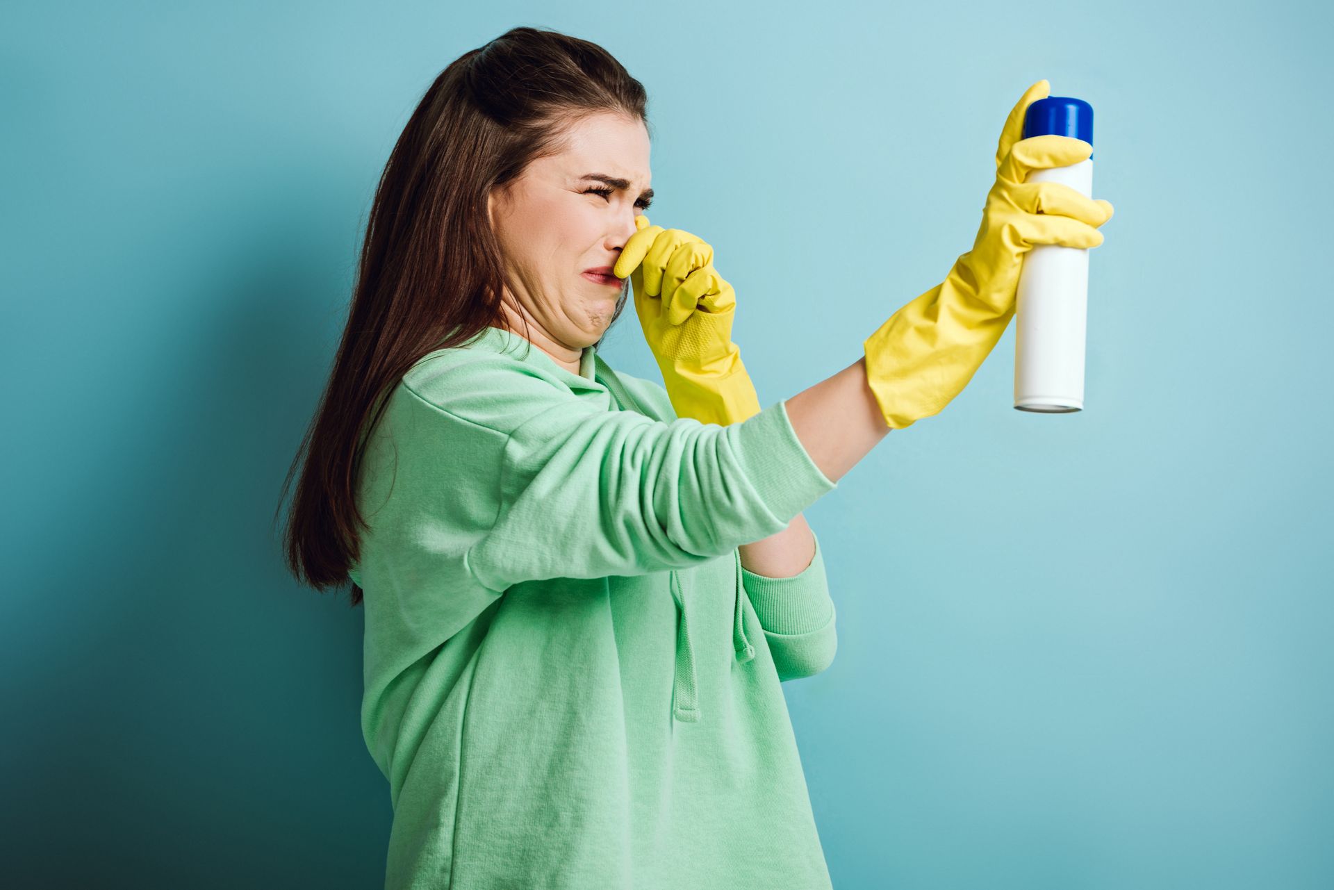 Person in green shirt and yellow gloves spraying disinfectant, holding nose against the smell.