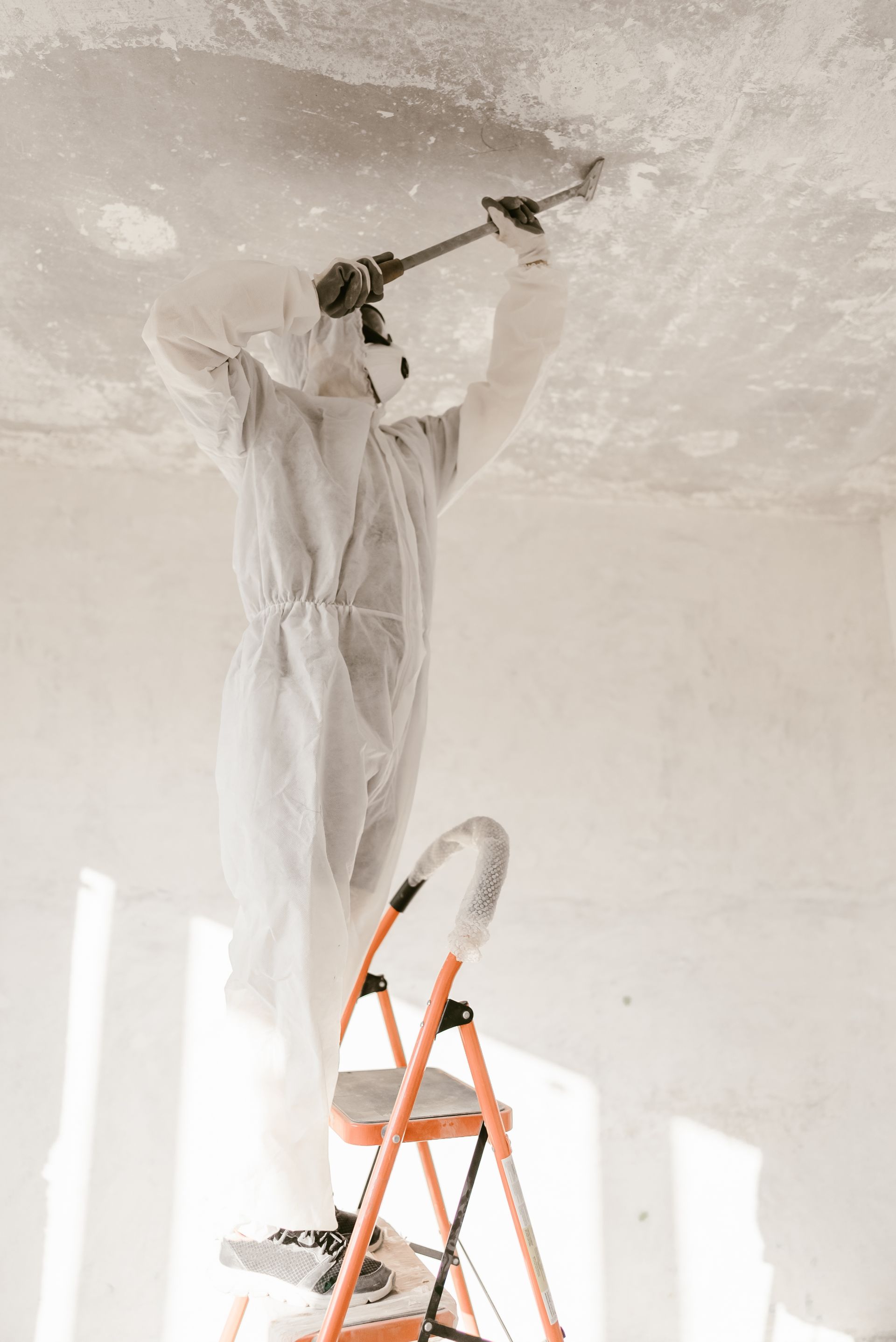 Person in white coveralls sanding a ceiling from a step ladder in a bright room