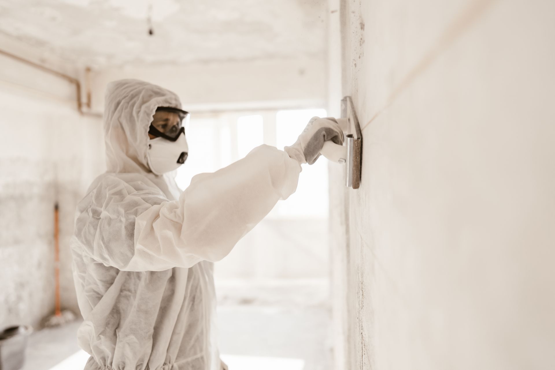 Person in white protective suit painting a wall with a roller, wearing a mask and goggles.