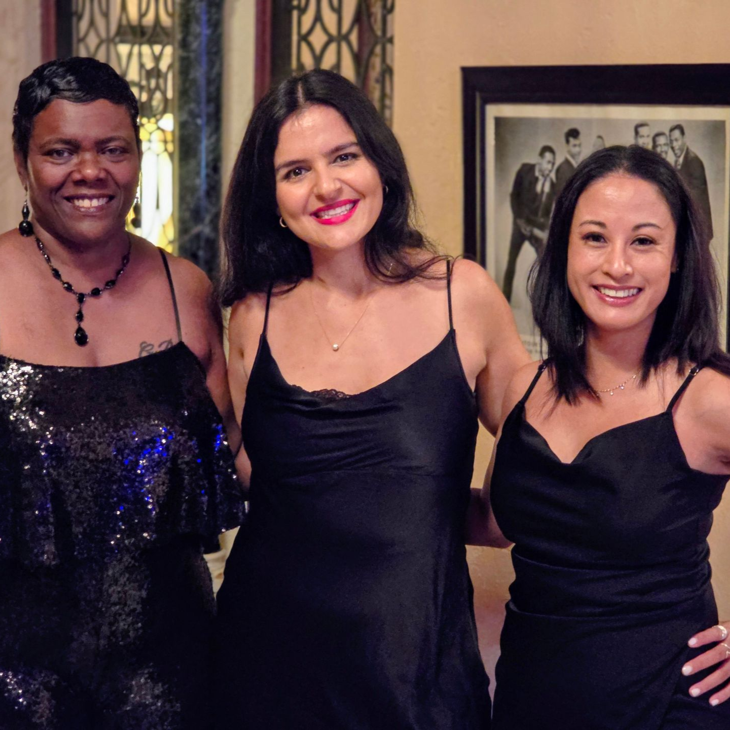 Three women smiling, dressed in black dresses, standing indoors.
