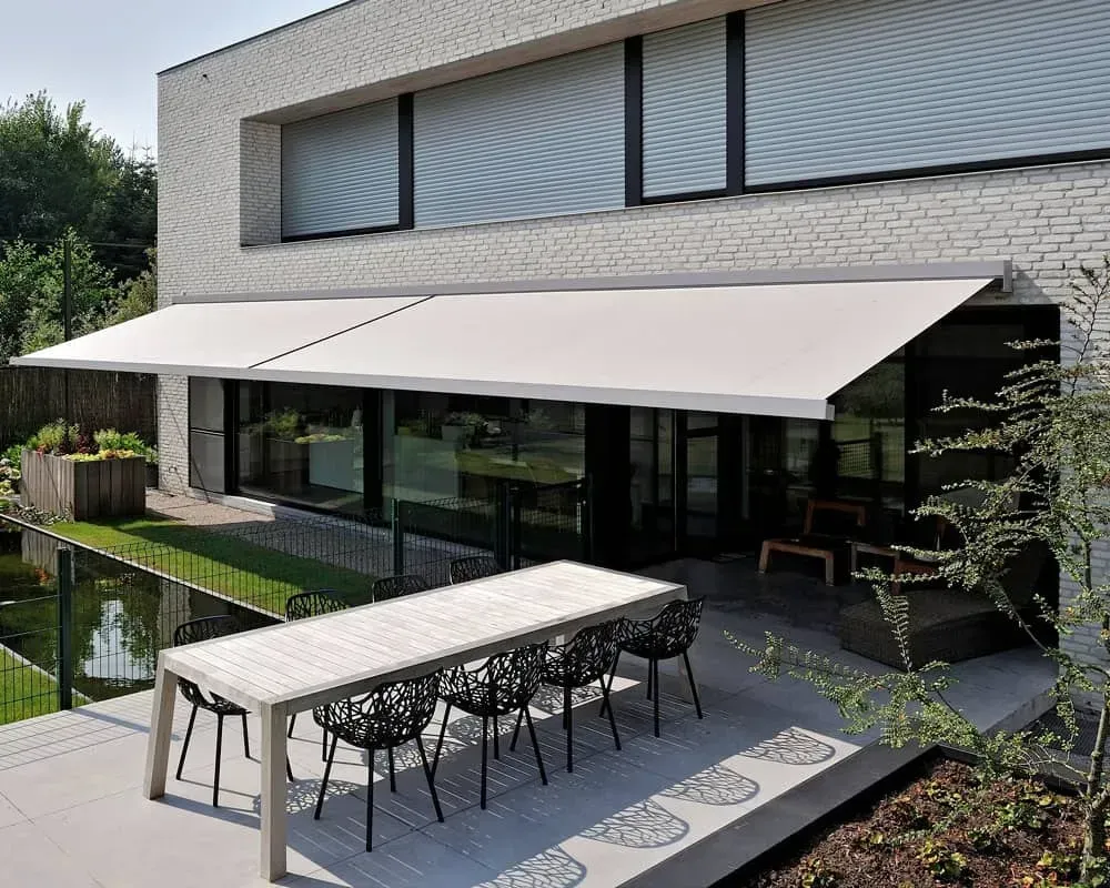 En udtrækkelig hvid markise skygger for en terrasse med spisebord og stole, nær et moderne hus med en spejlbassin.