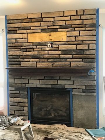Stone fireplace under renovation with a dark mantel, blue tape on the right edge, and tools on the floor.