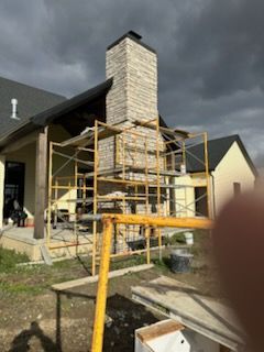 Stone chimney under construction with yellow scaffolding beside a house, under dark cloudy skies