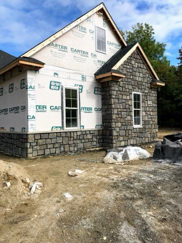 Partially built house with stone chimney, wrapped in house sheathing, on a dirt lot under a blue sky