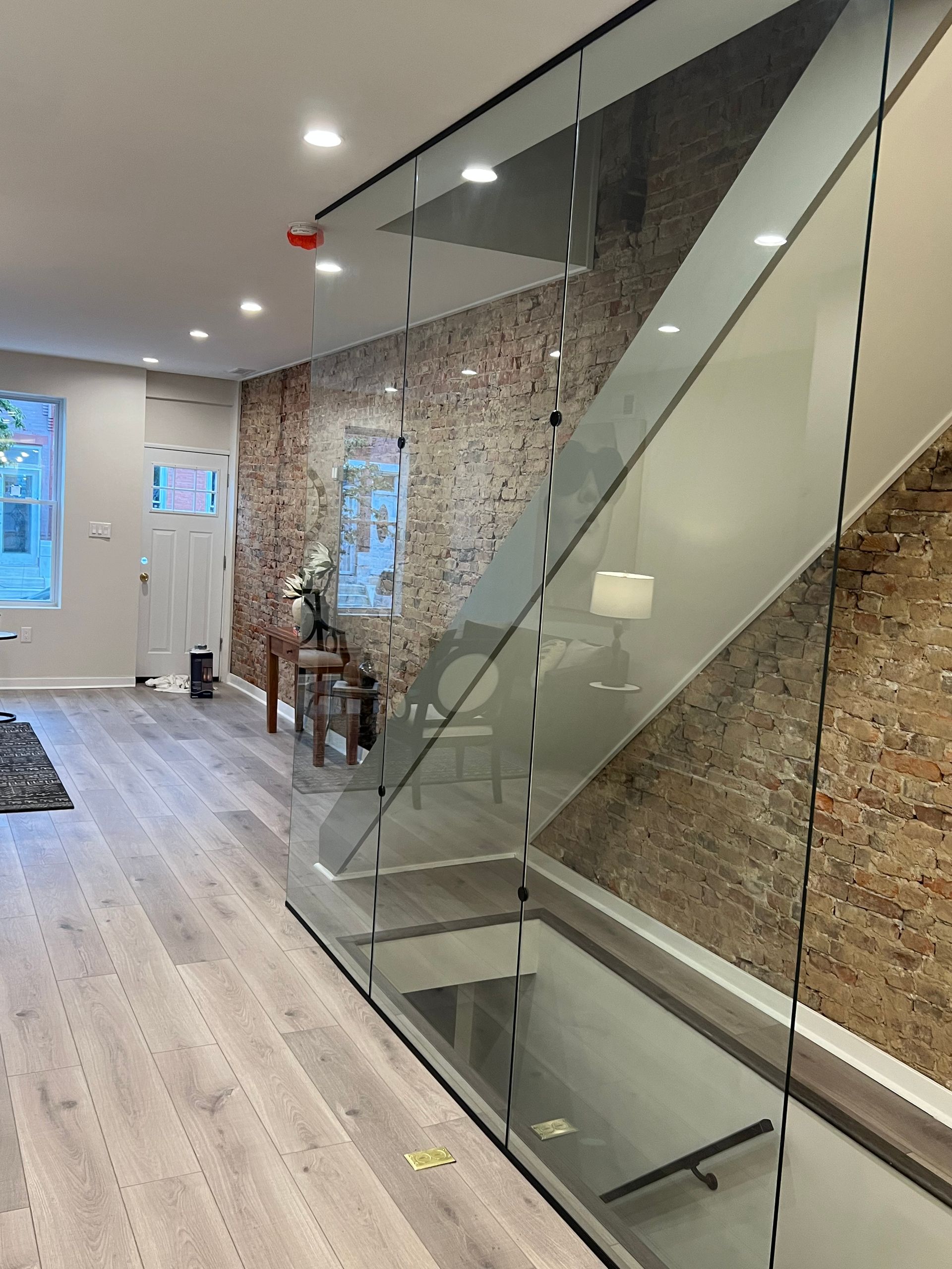 A modern room with light wood floors, a brick feature wall, a glass staircase railing, and recessed ceiling lighting.